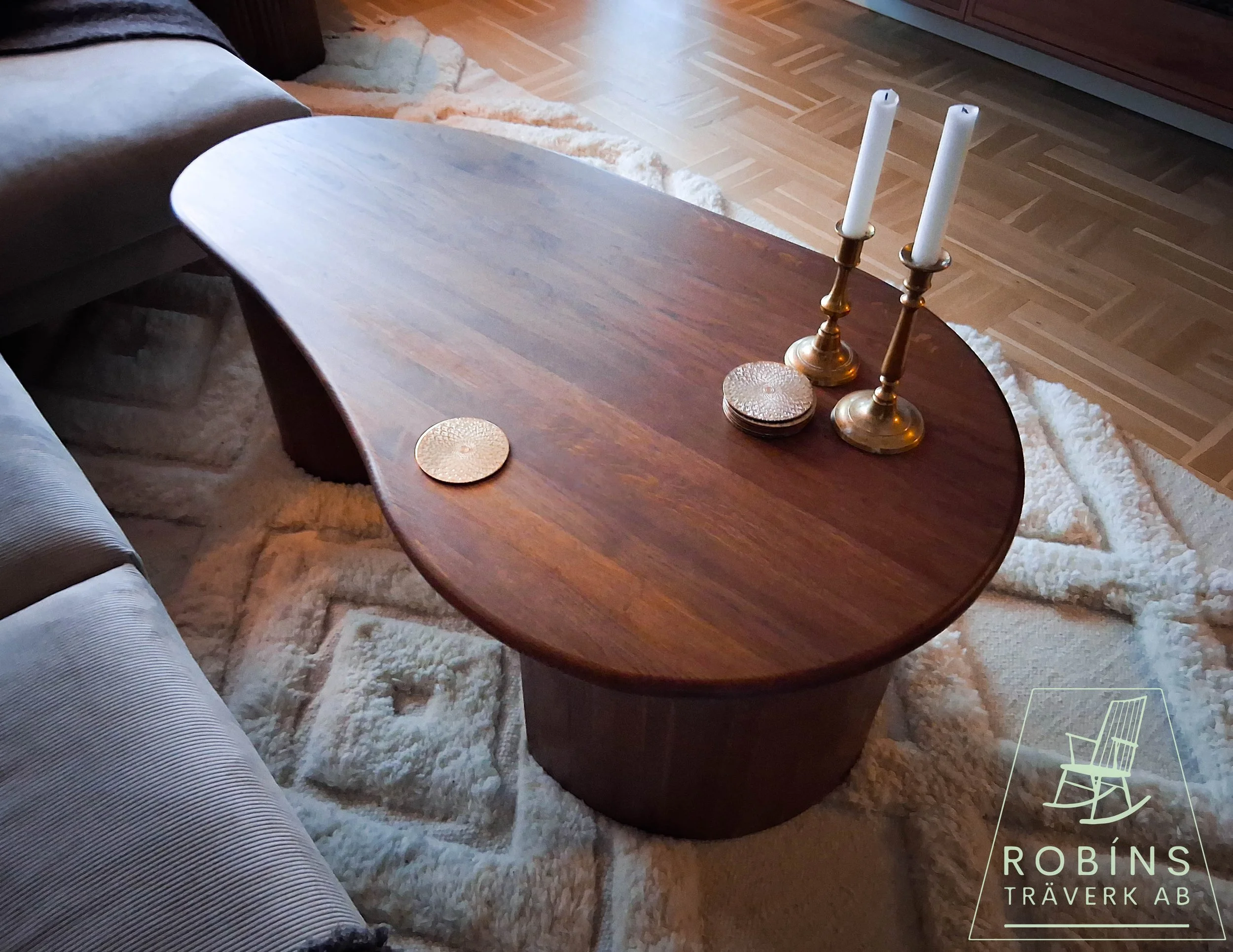A wooden coffee table with a polished surface, placed on a cream-colored shaggy rug in a living room. The table has a round gold-colored decorative item, two gold candlestick holders with white candles, and a small round decorative bowl. Part of a so