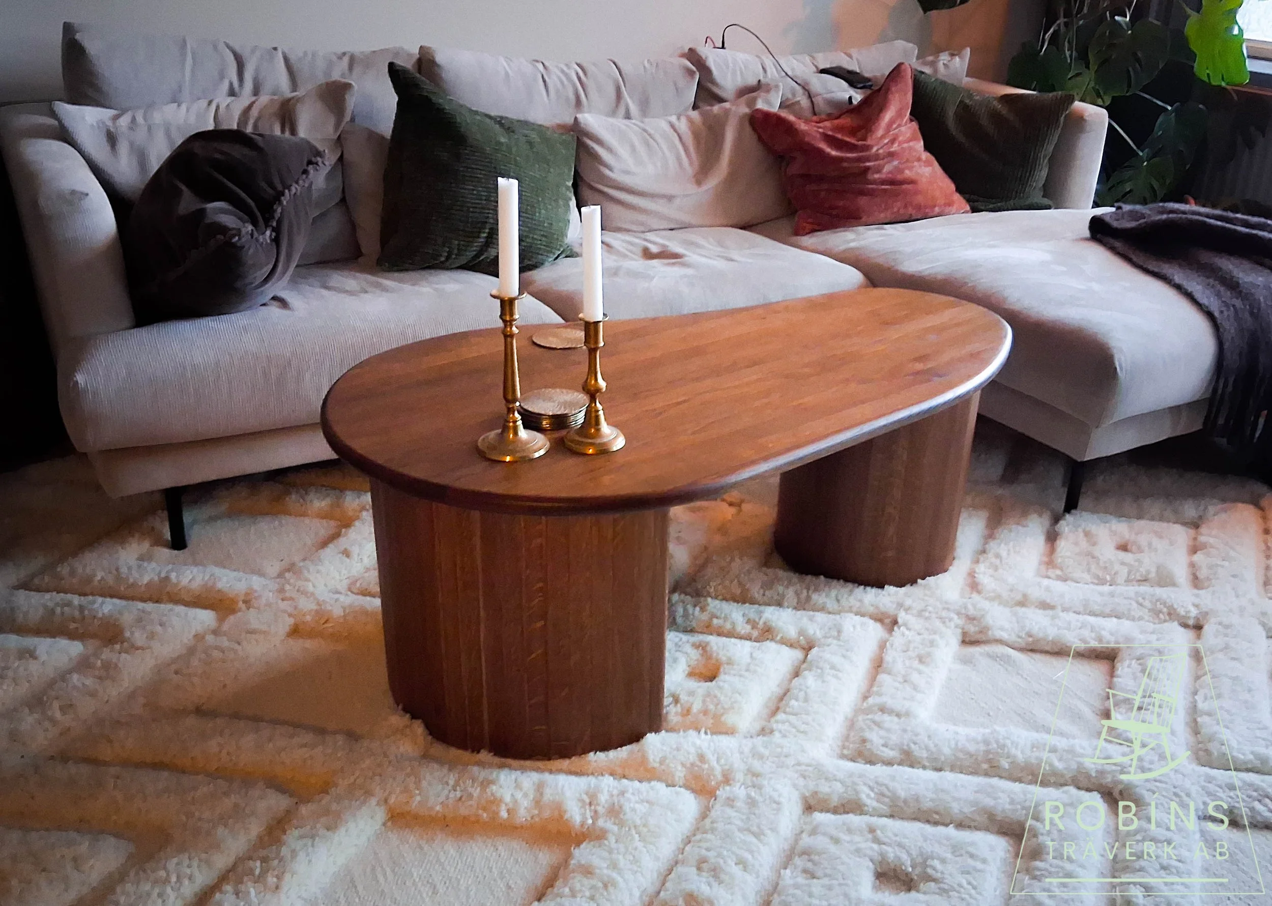 A living room features a beige sectional sofa with multiple throw pillows, a wooden oval coffee table with two brass candlesticks and candles, a cream-colored textured rug, and potted plants near the window.