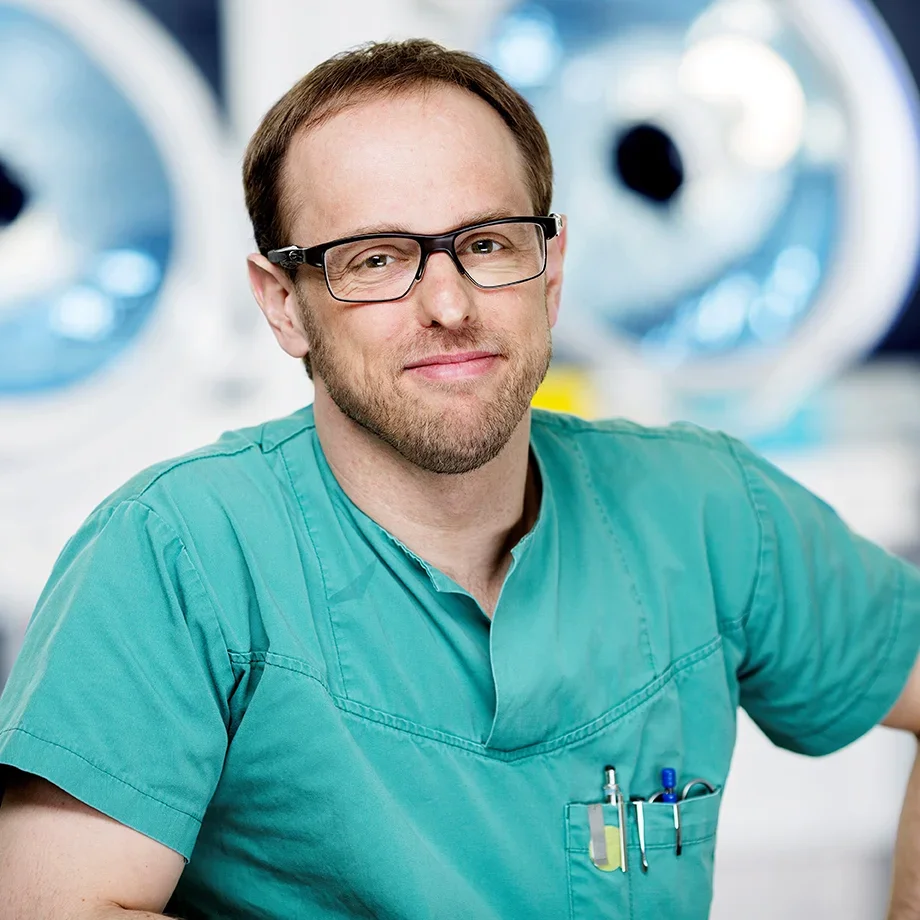 En mann iført kirurgisk uniform og briller, som smiler, med en blåmaskin i bakgrunnen.
