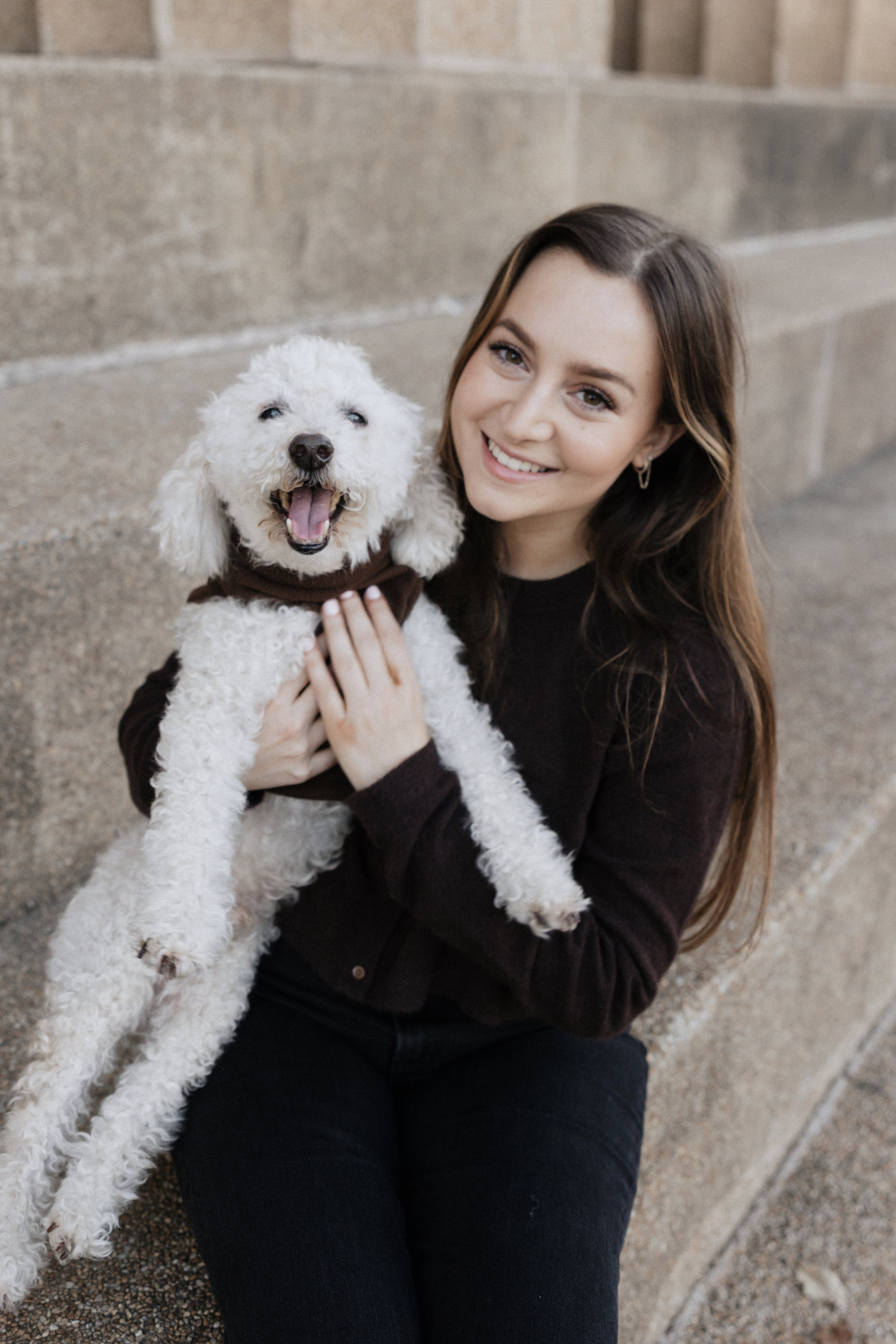 A woman with long brown hair smiling while holding a white fluffy dog.