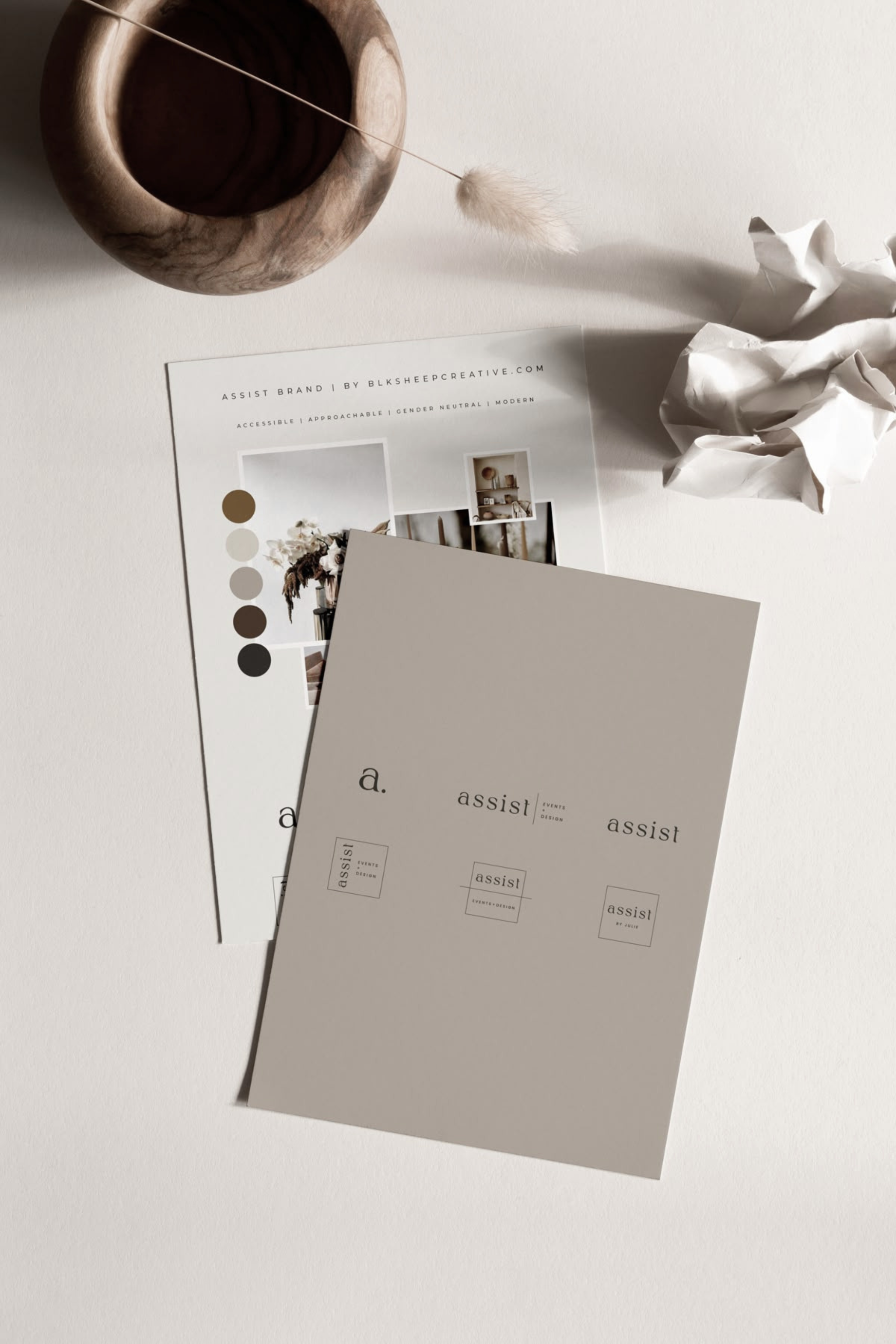 A flat lay of minimalist office or branding materials, including monochrome color palettes, a brochure, and a crumpled sheet of paper, on a white surface with a wooden bowl and a fluffy decorative object.