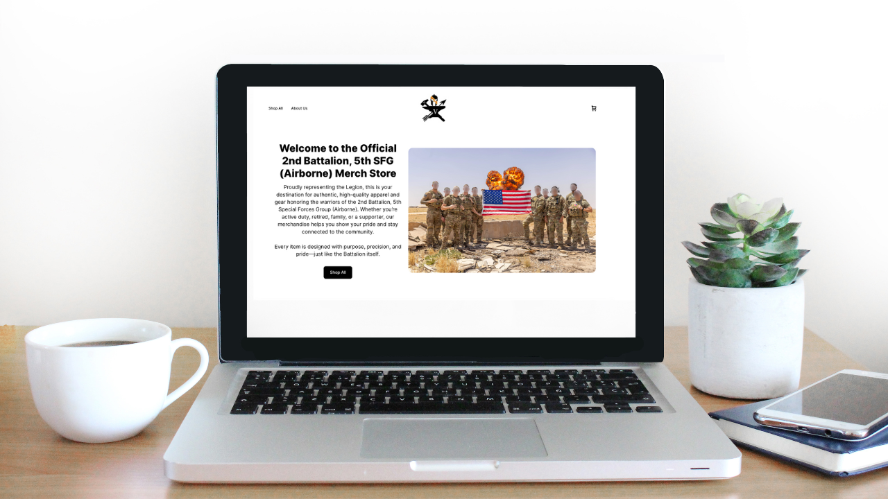 Laptop displaying a website about the 2nd Battalion, 5th SFG (Airborne) merchandise store on a wooden desk with a white coffee mug, a potted succulent plant, and a closed notebook with a smartphone on top.