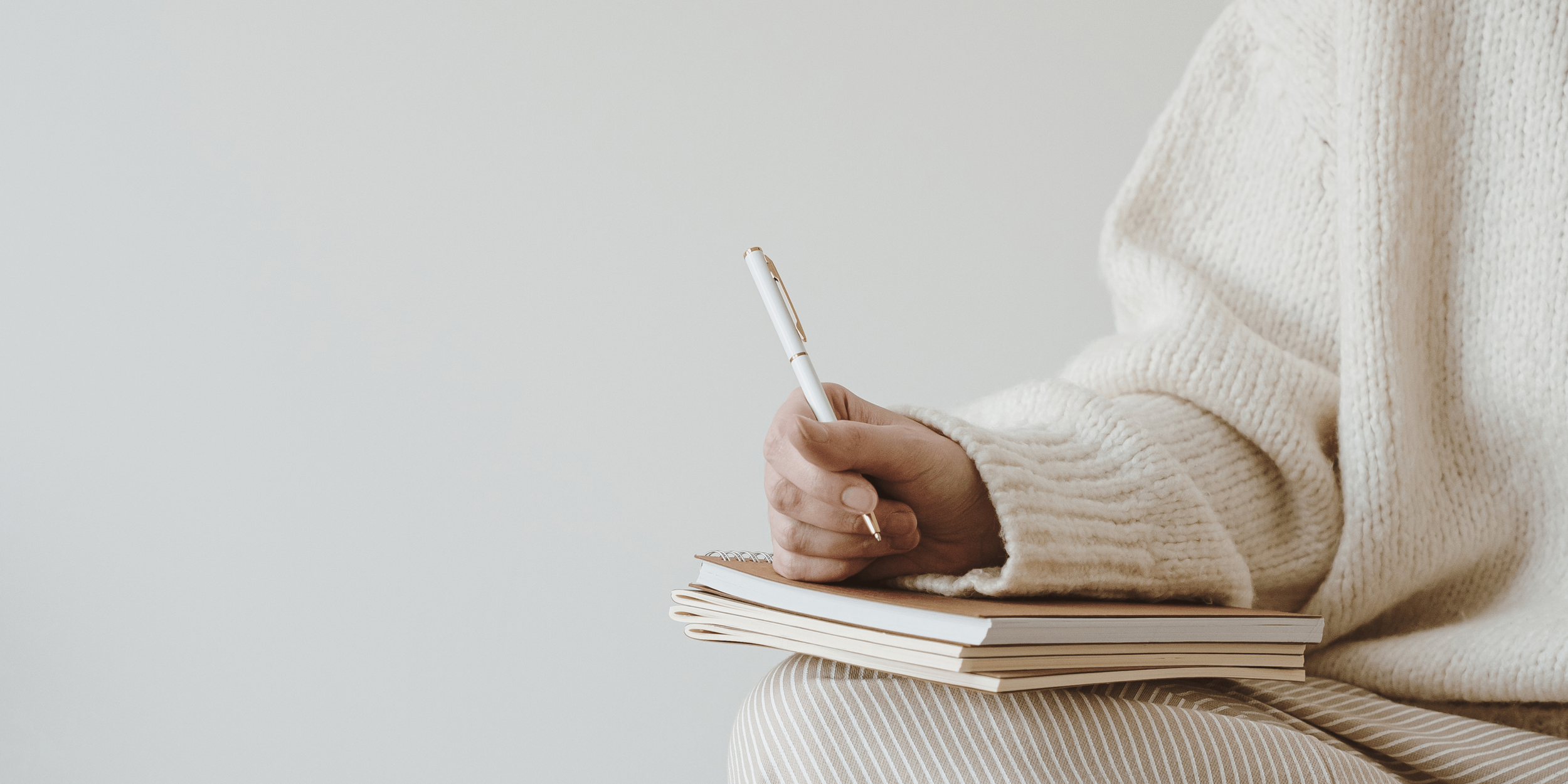 Person wearing a cream sweater holding a pen, sitting with a stack of notebooks