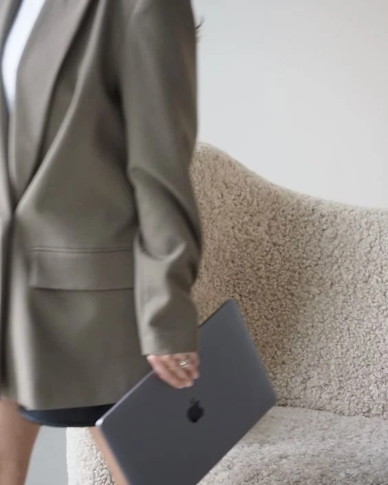 Close-up of a person in a gray blazer holding a closed MacBook laptop, standing next to a beige textured armchair.