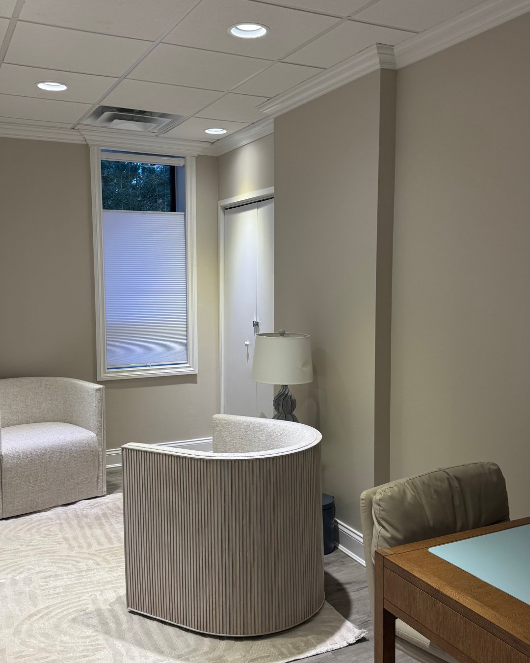 A corner of an indoor room showing a beige couch, a curved ribbed side table, a white lamp on a stand, a window with a partially closed blind, a door, and a ceiling with recessed lights.
