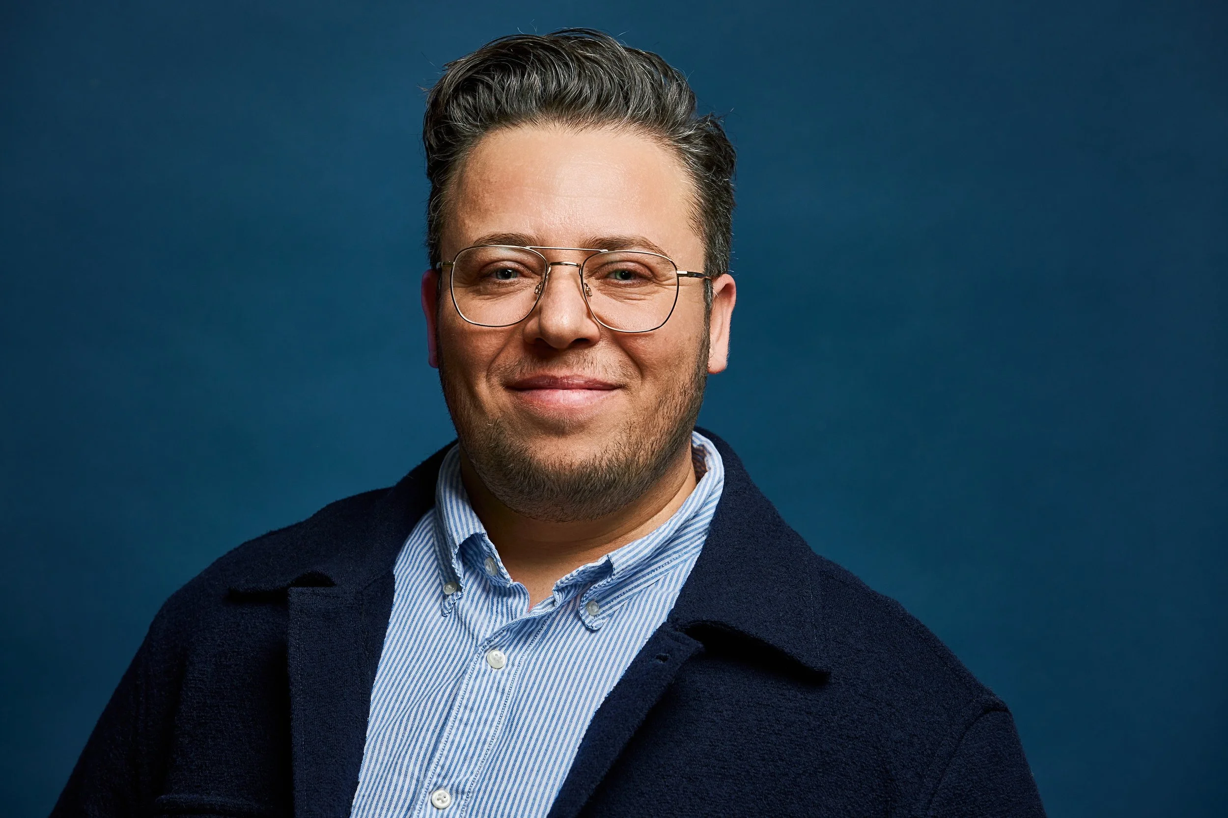 Portrait of a smiling man with glasses, dark hair, wearing a light blue dress shirt and a dark navy sweater, against a dark blue background.