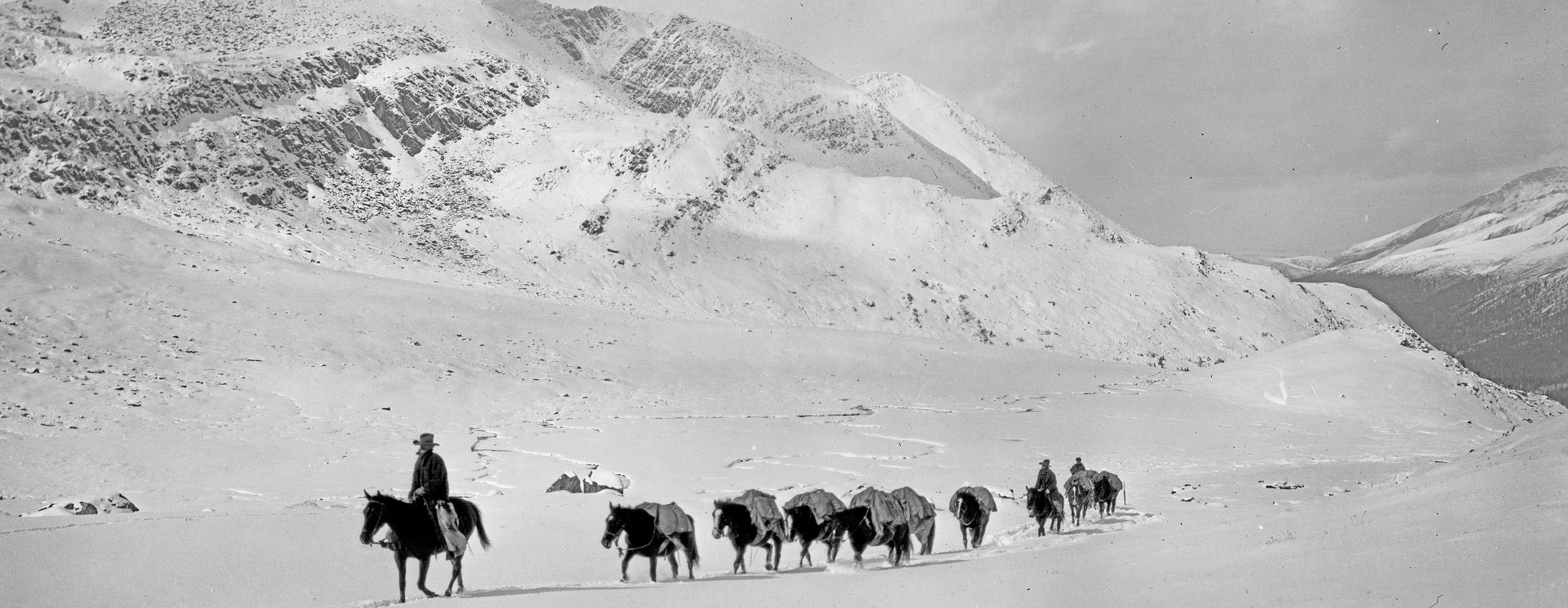 A black and white photograph of a snow-covered mountainous landscape with seven horse riders traveling single file, some carrying supplies, under a partly cloudy sky.