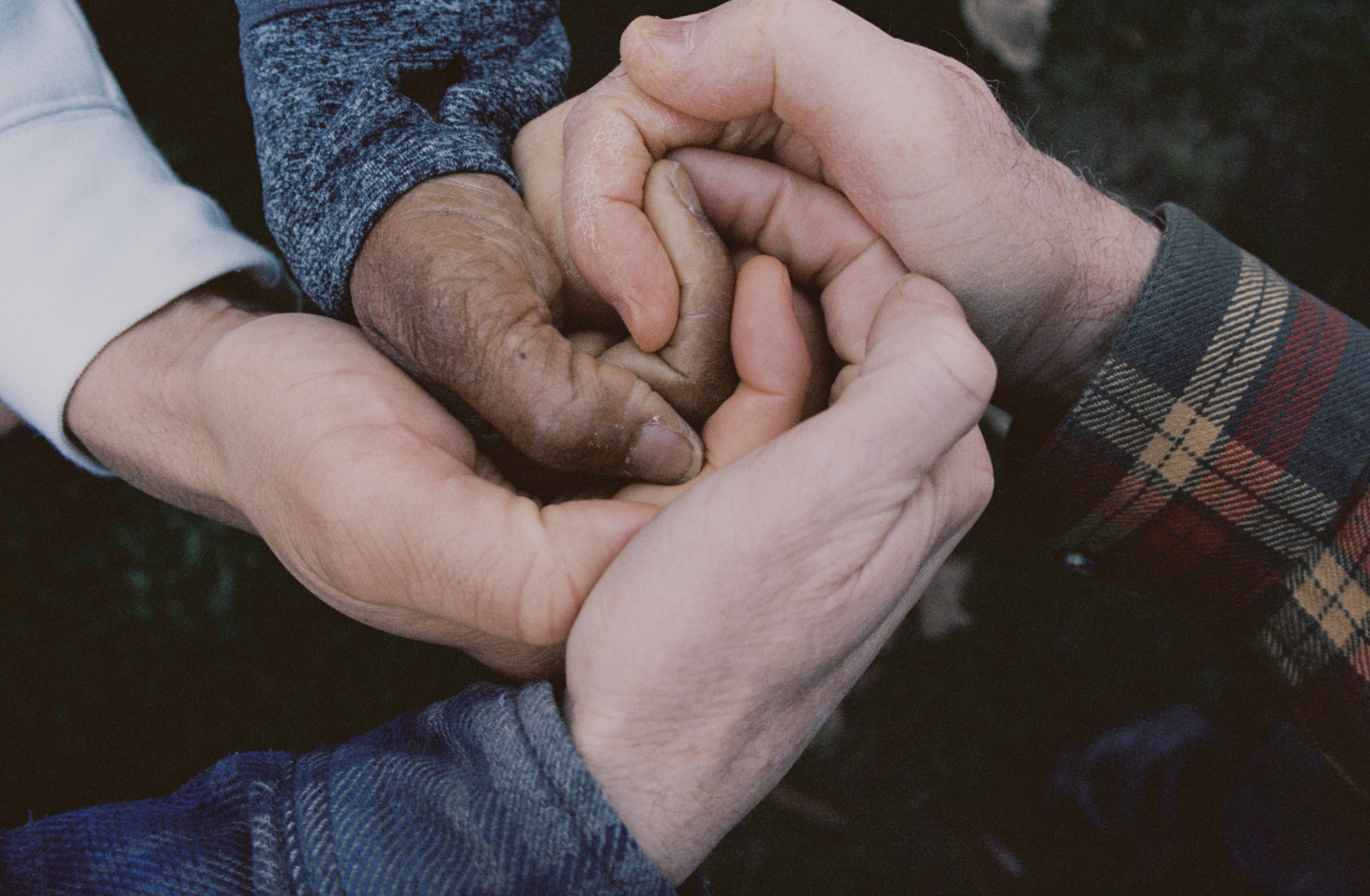 Multiple hands, including a darker-skinned hand, holding and interlocking with lighter-skinned hands in a caring manner.