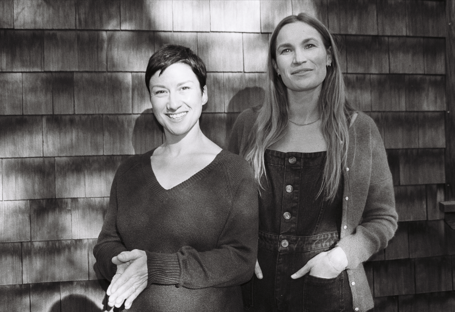Two women standing side by side in front of a wooden wall, smiling and looking at the camera. The woman on the left has short dark hair and is wearing a dark sweater, while the woman on the right has long, wavy hair and is wearing a dark denim dress with a cardigan.