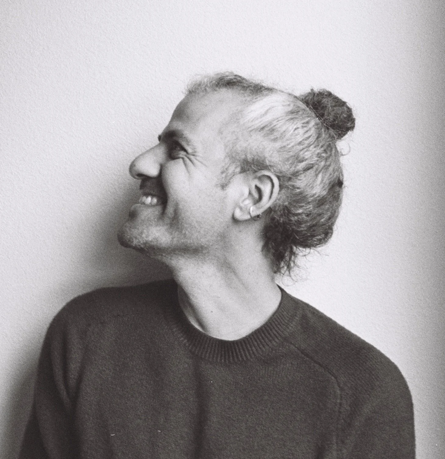 Side profile of a smiling man with long hair tied in a bun, wearing a dark sweater, against a plain wall.