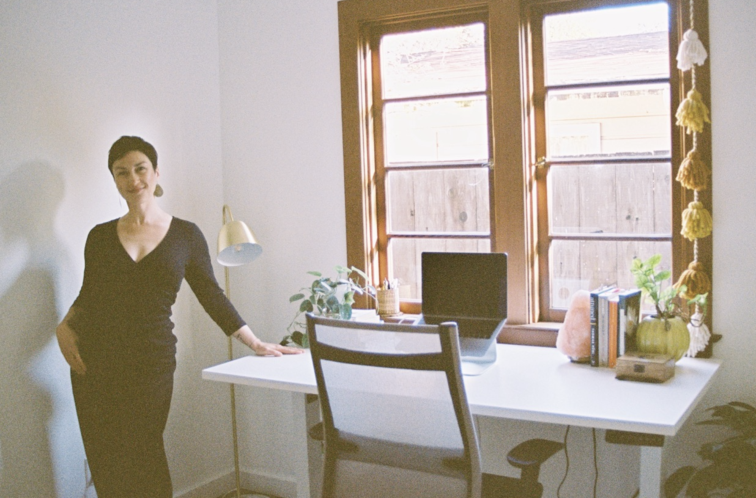 Woman in black dress standing by white desk near window in room with white walls and wooden window frames
