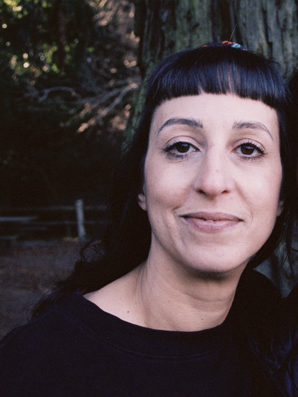 A woman with black hair and bangs, smiling slightly, outdoors near a large tree.