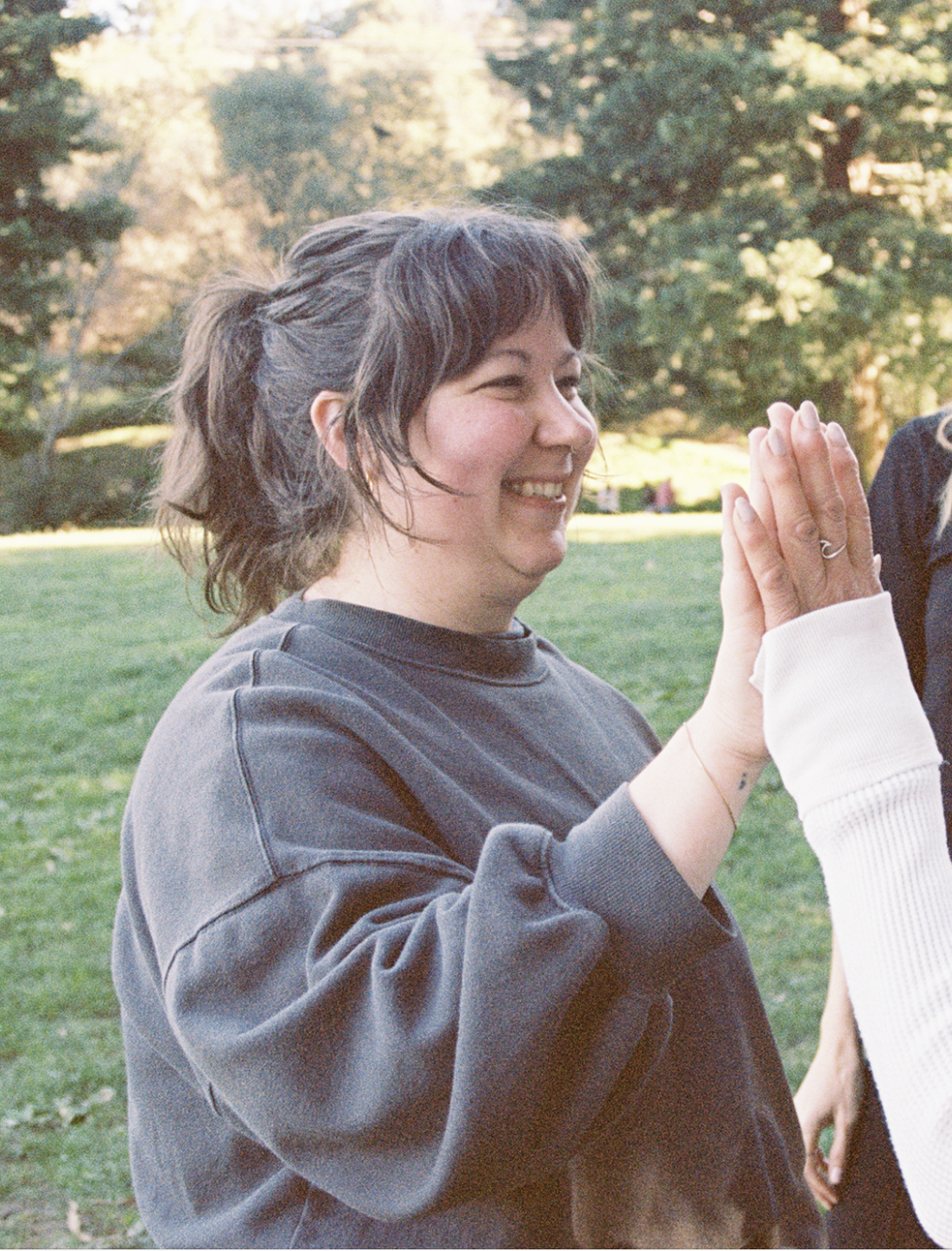 A woman with short brown hair smiling and giving a high-five outdoors in a park with trees in the background.