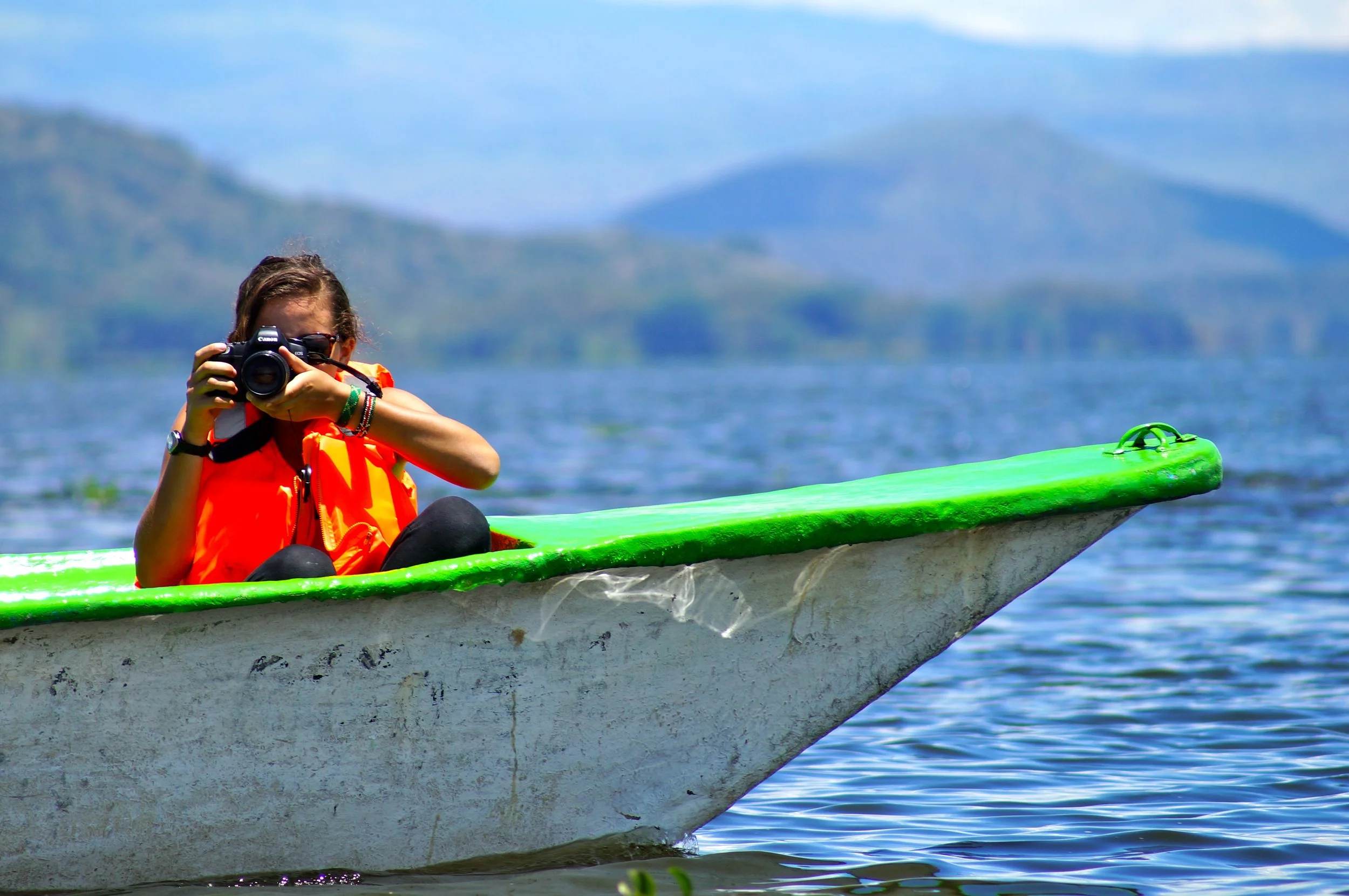 A young woman in an orange life jacket sitting on a small boat on a lake, holding a camera up to her face and taking a photo. In the background, there are hills and mountain ranges under a blue sky.