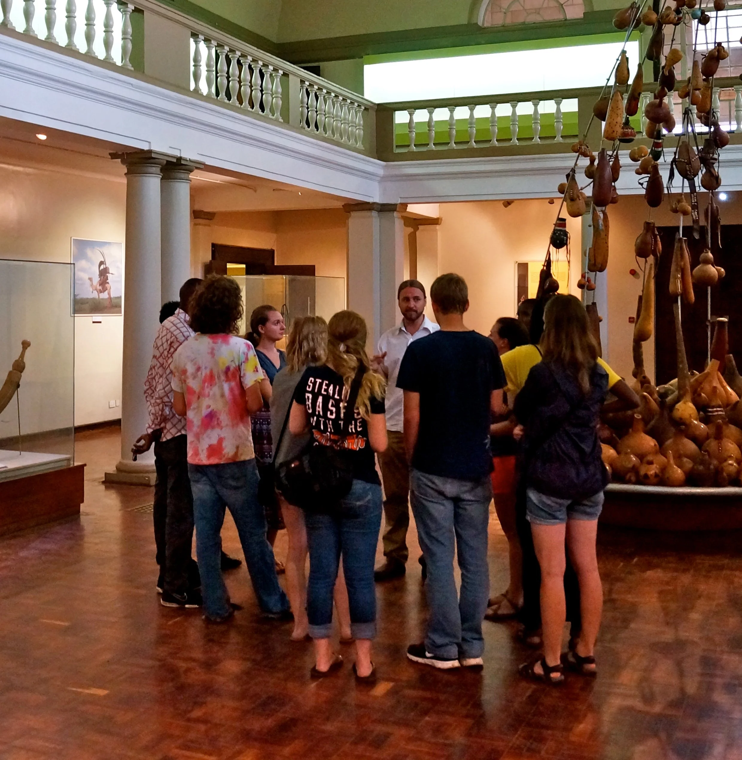 Group of people standing in a museum exhibition area, listening to a guide. The room has a large hanging sculpture made of gourds and a painting on the wall.