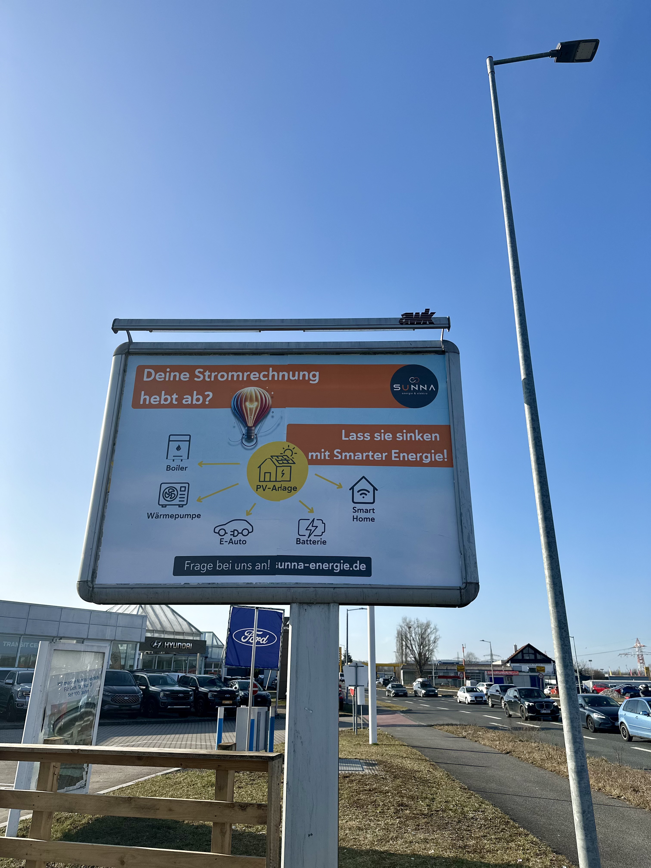 Werbeplakat für SUNNA Energie mit Informationen zu intelligenter Energieverwendung wie PV-Anlagen, Wärmepumpen, E-Autos, Batterien und Smart Home. Slogan: 'Deine Stromrechnung hebt ab? Lass sie sinken mit Smarter Energie!' Standort an einer Straße mi