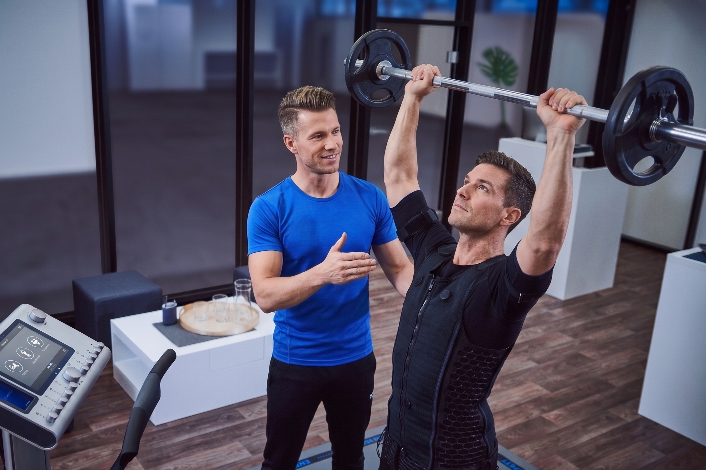 Ein Mann steht auf einem Laufband im Fitnessstudio und hebt eine Hantel, während eine Trainerin ihm Ratschläge gibt.