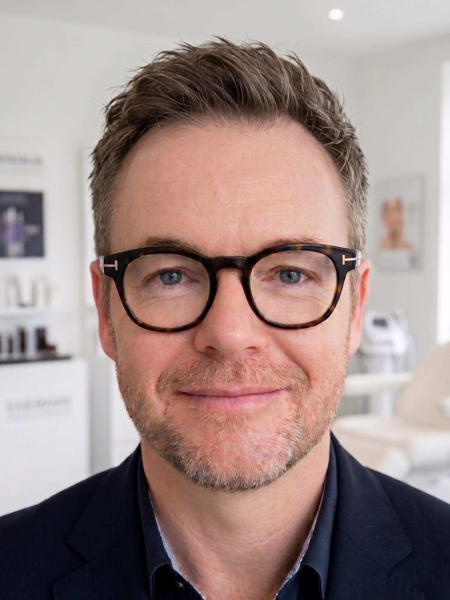 Nahaufnahme eines Mannes mit braunen, gelockten Haaren und blauen Augen, trägt schwarze Brillen und ein dunkles Jackett, im Hintergrund ein helles, modernes Raum mit Bildern an der Wand.
