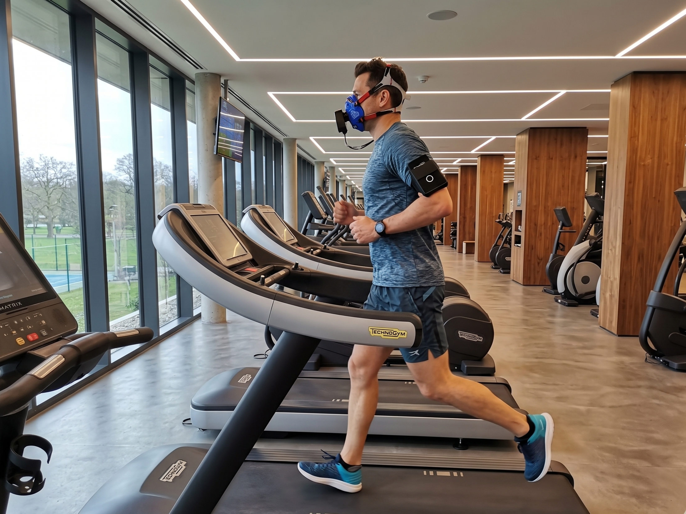 Mann beim Lauftraining auf einem Laufband im Fitnessstudio, trägt eine blauen Maske, Sportbekleidung und einen Pulsmesser am Oberarm, im Hintergrund große Fenster mit Blick ins Freie.