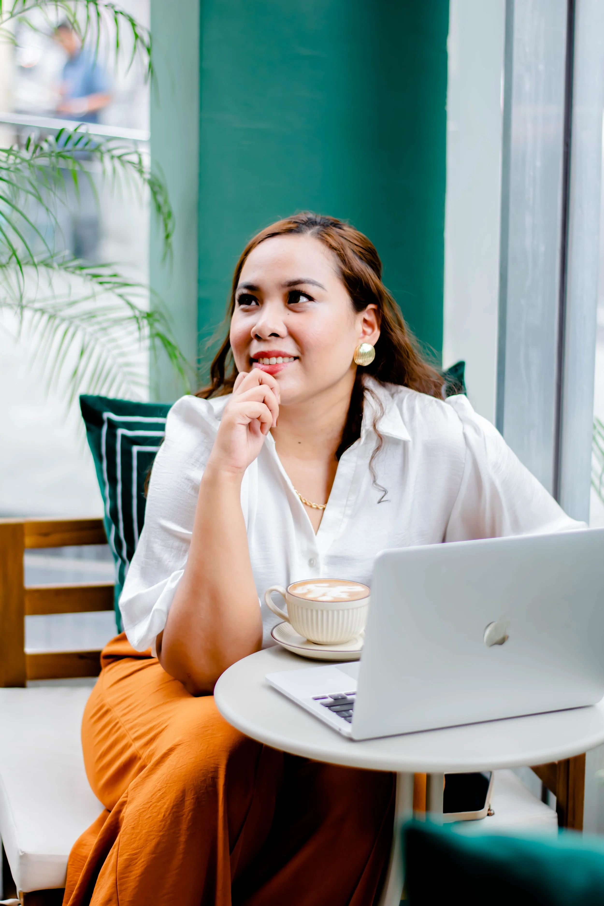 A woman with brown hair and gold earrings, sitting at a white round table with a white laptop and a cup of coffee, smiling and looking to the side in a cafe with green walls and plants.