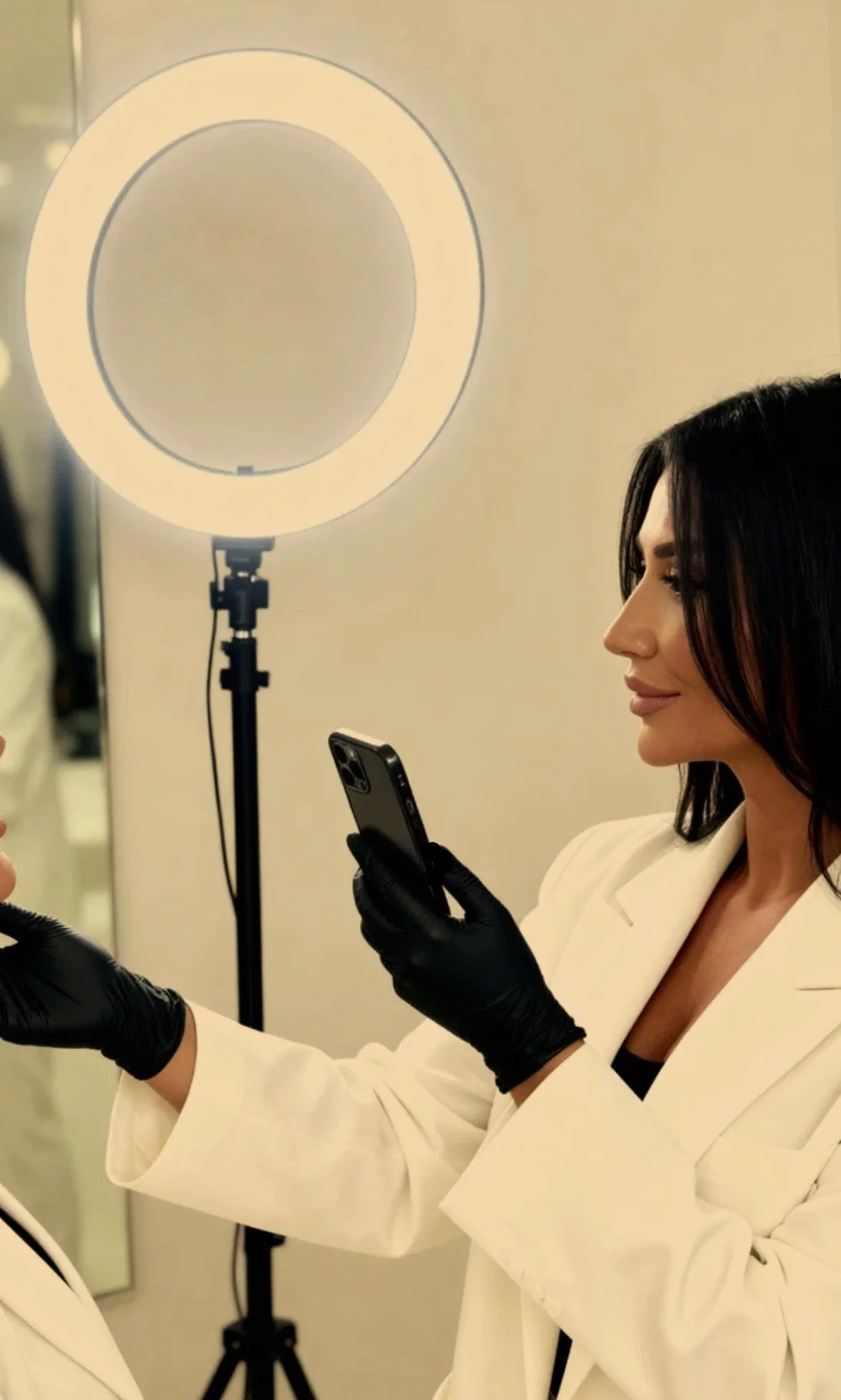 Frau in Weißer Jacke und schwarzen Handschuhen hält ein Smartphone und posiert vor einem Ringlicht.