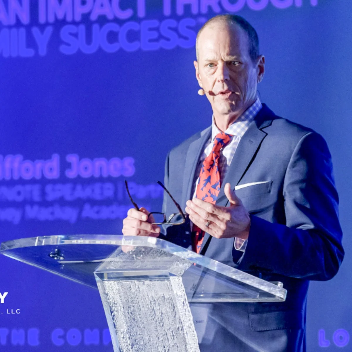 A man in a blue suit and patterned tie speaking at a podium with a purple background with partially visible text.