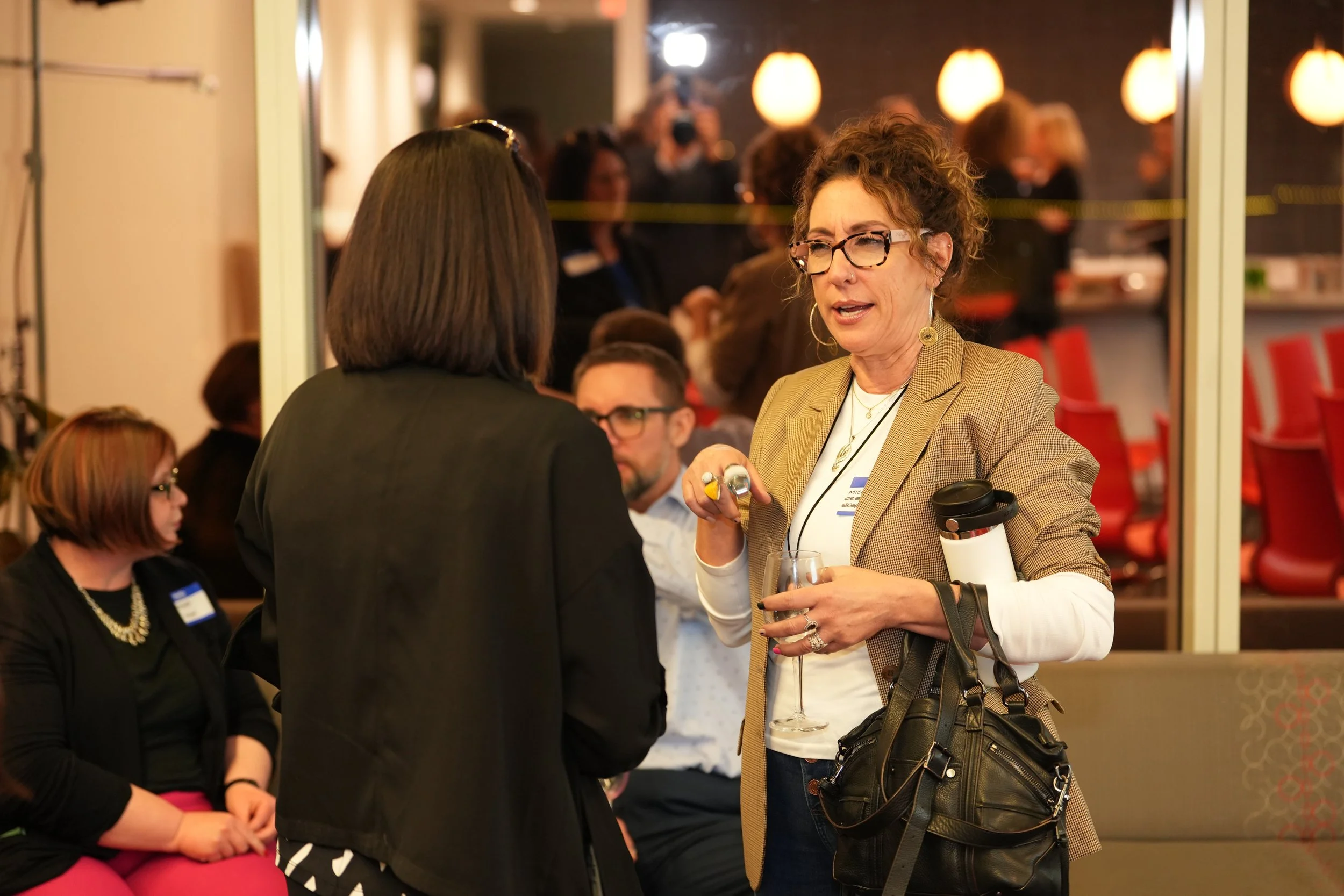 People at a networking event, one woman holding a drink, talking to another woman, with others seated and standing in the background.