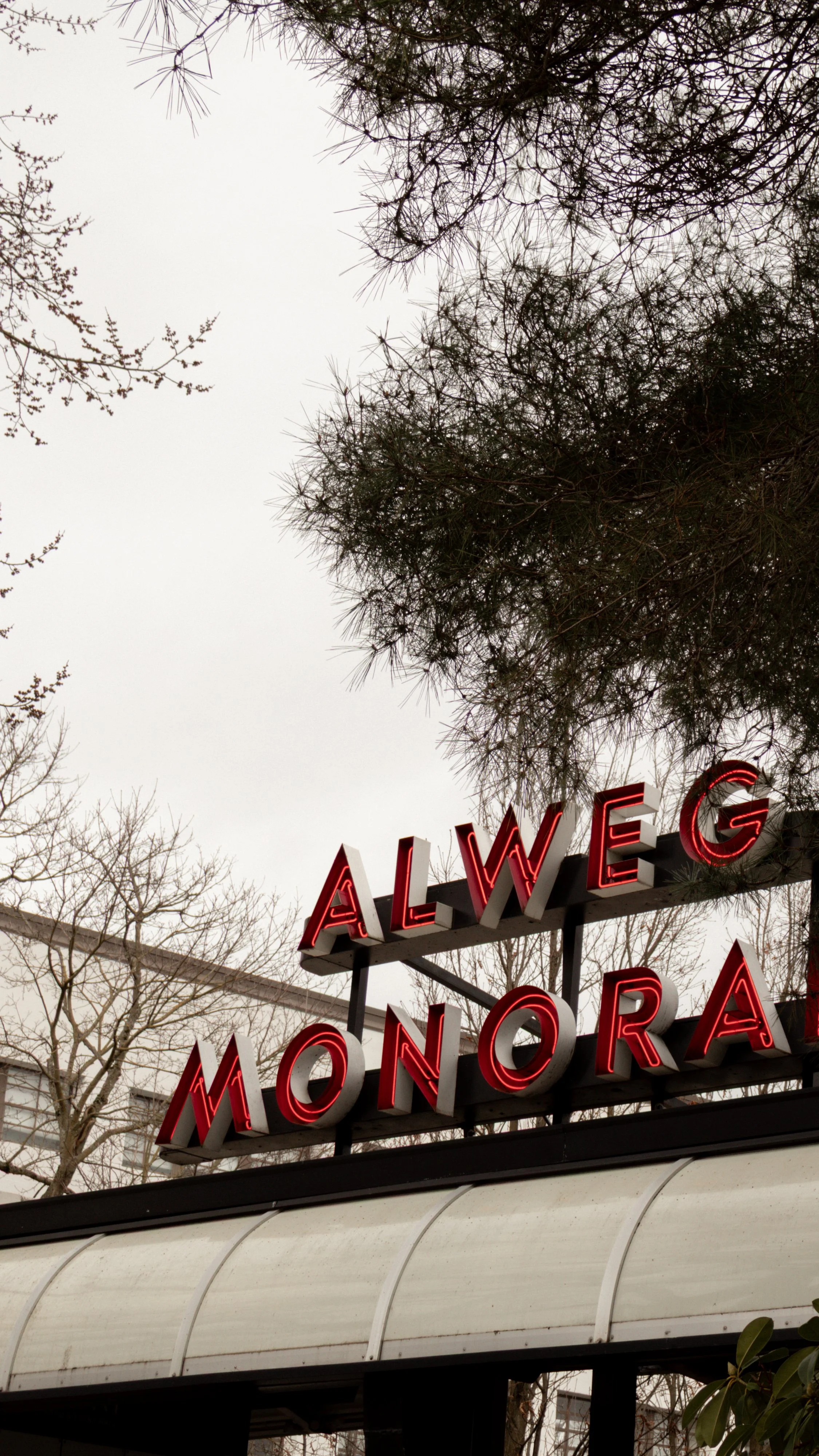 Red neon sign reading 'ALWEG MONORA' above an entrance, with trees and an overcast sky in the background.