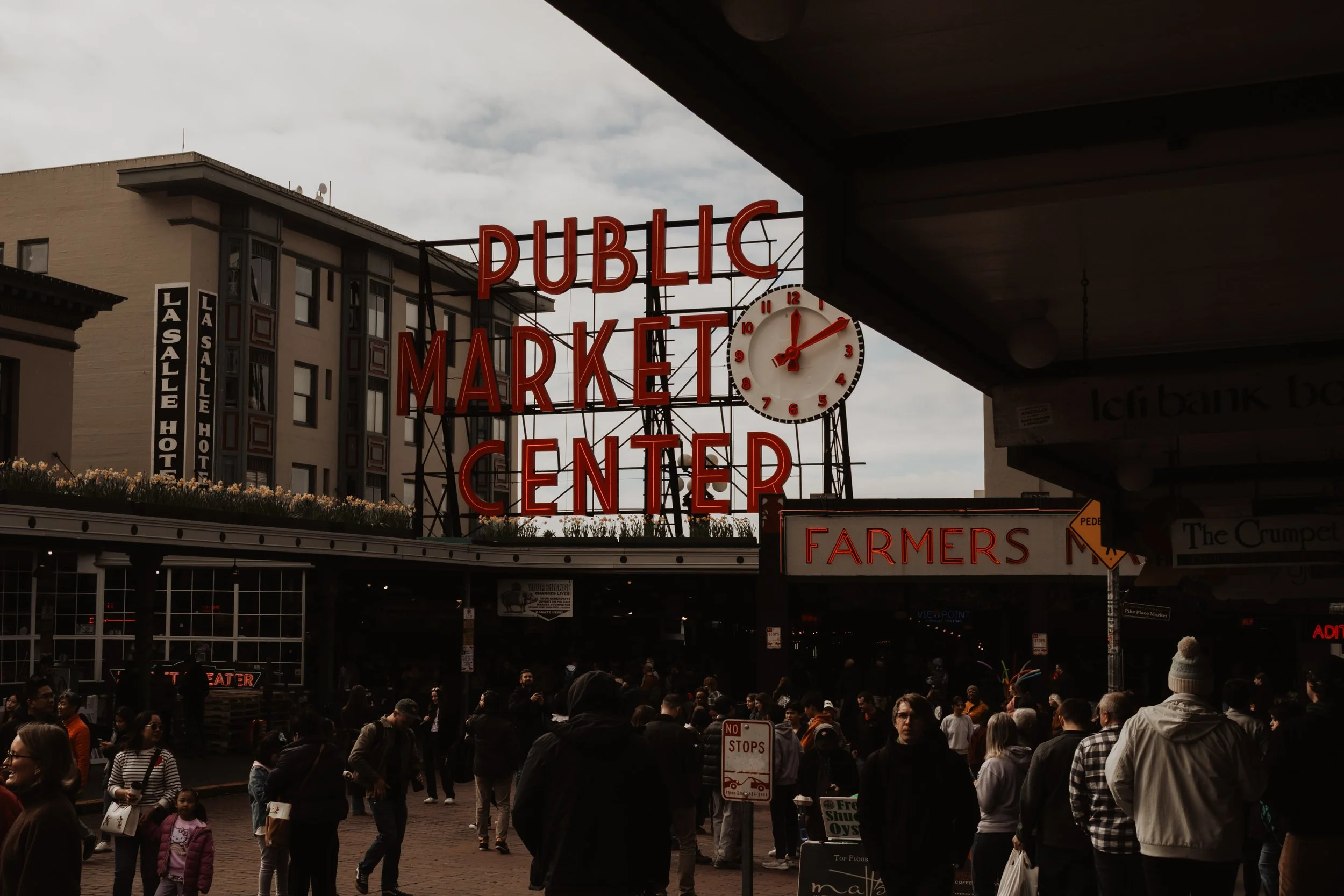 The entrance to Pike Place Market in Seattle with a sign reading 'Public Market Center' and a clock showing 3:03, amidst a busy crowd of people.