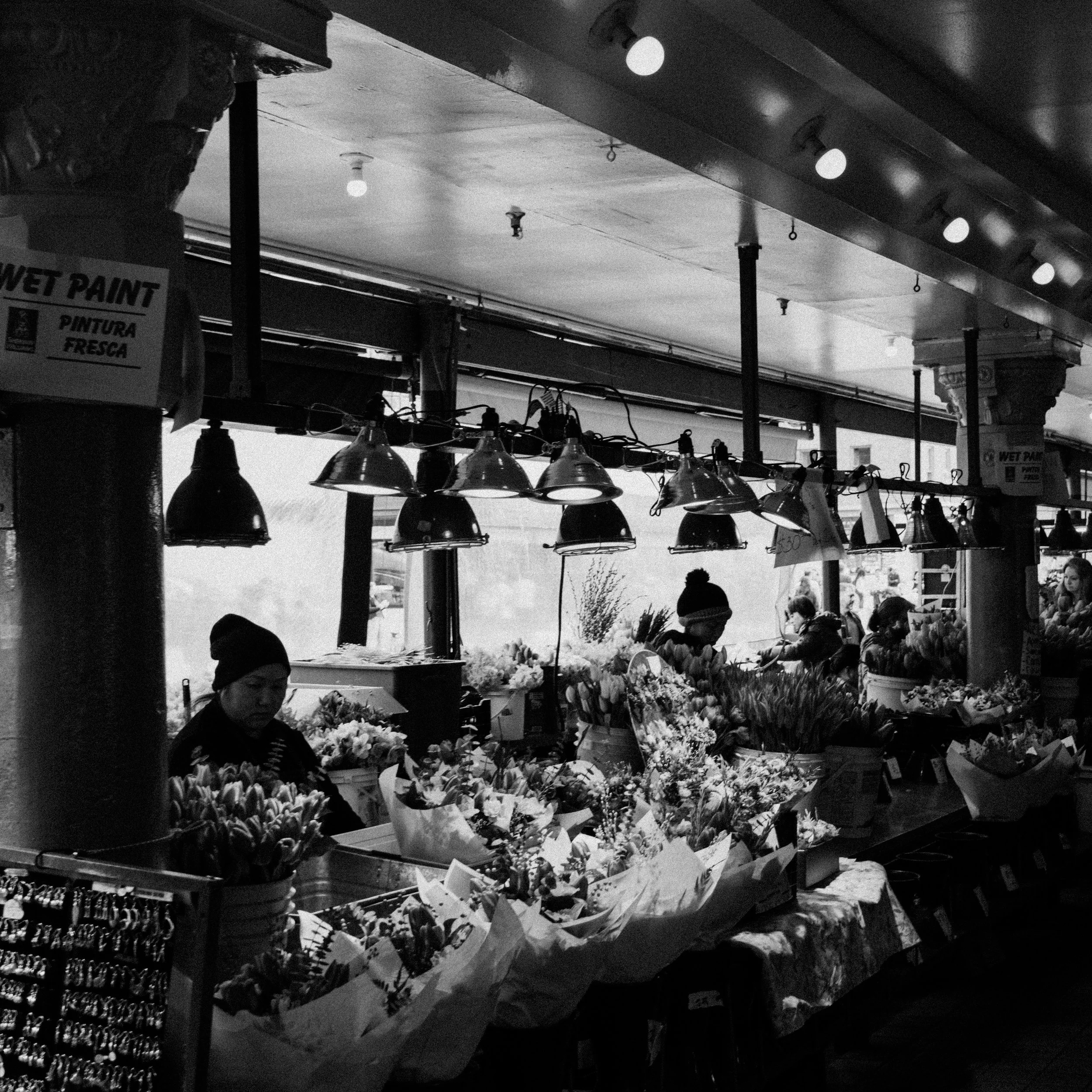 A black-and-white photo of a flower stall at a market, with three people arranging flowers behind the counter. The stall has overhead lighting and signs advertising wet paint in English and Spanish.