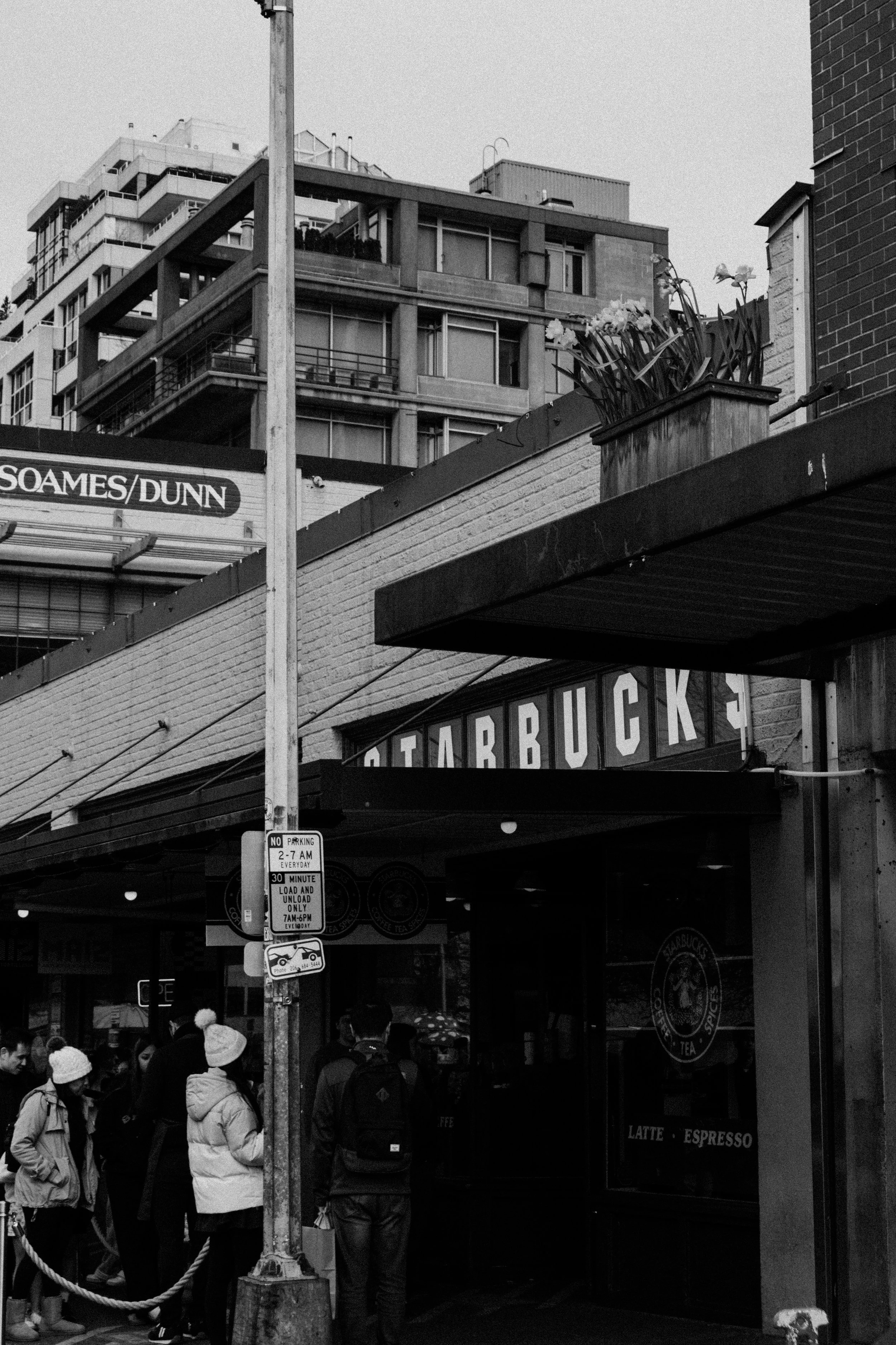 Black and white photo of a bustling street scene outside a Starbucks coffee shop with people waiting in line. The Starbucks sign is visible, along with a street pole and signs, including one indicating parking restrictions. Residential buildings are 
