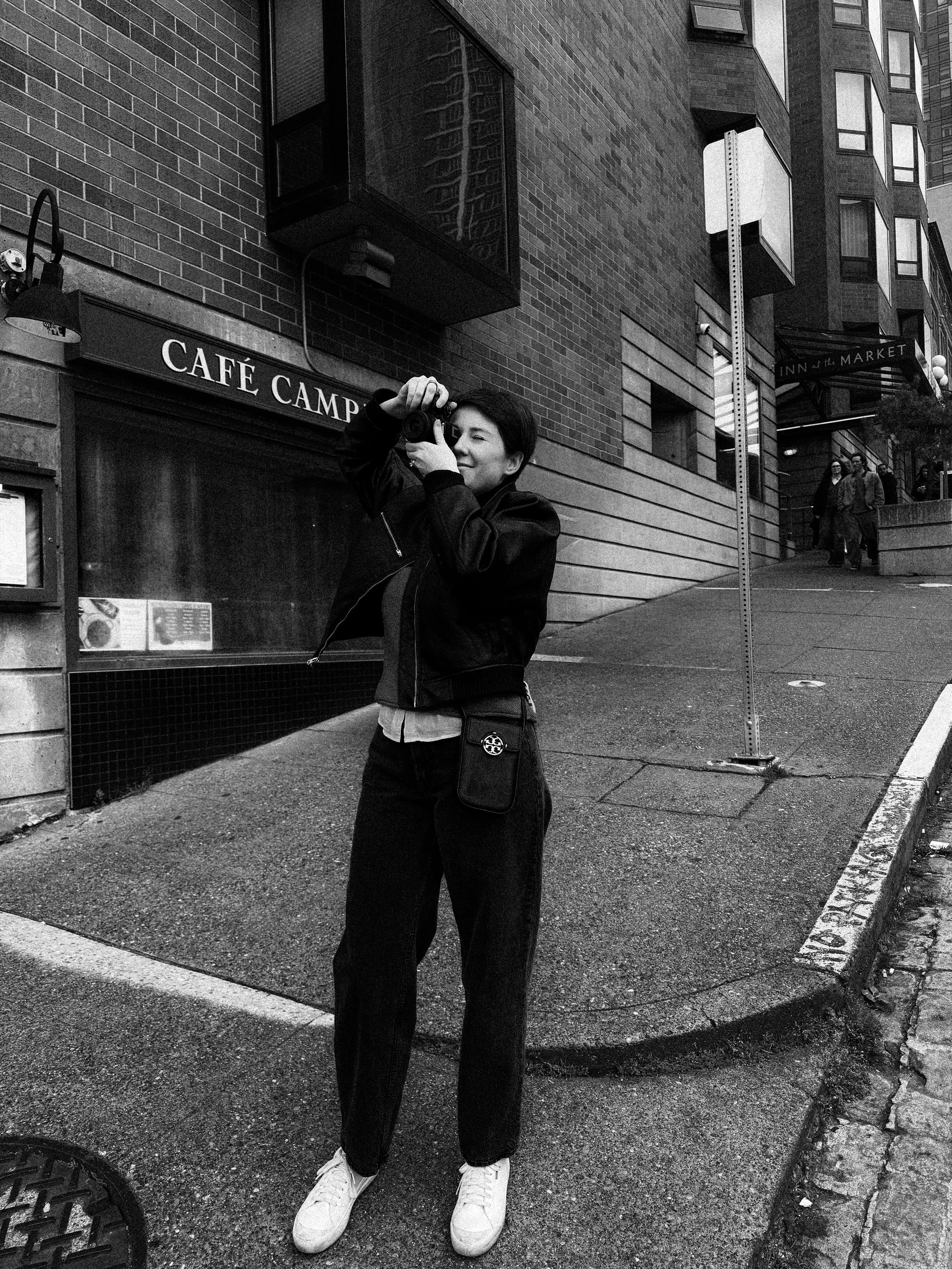 A person standing on a city sidewalk, taking a photograph with a camera, near a café called 'Café Camp'