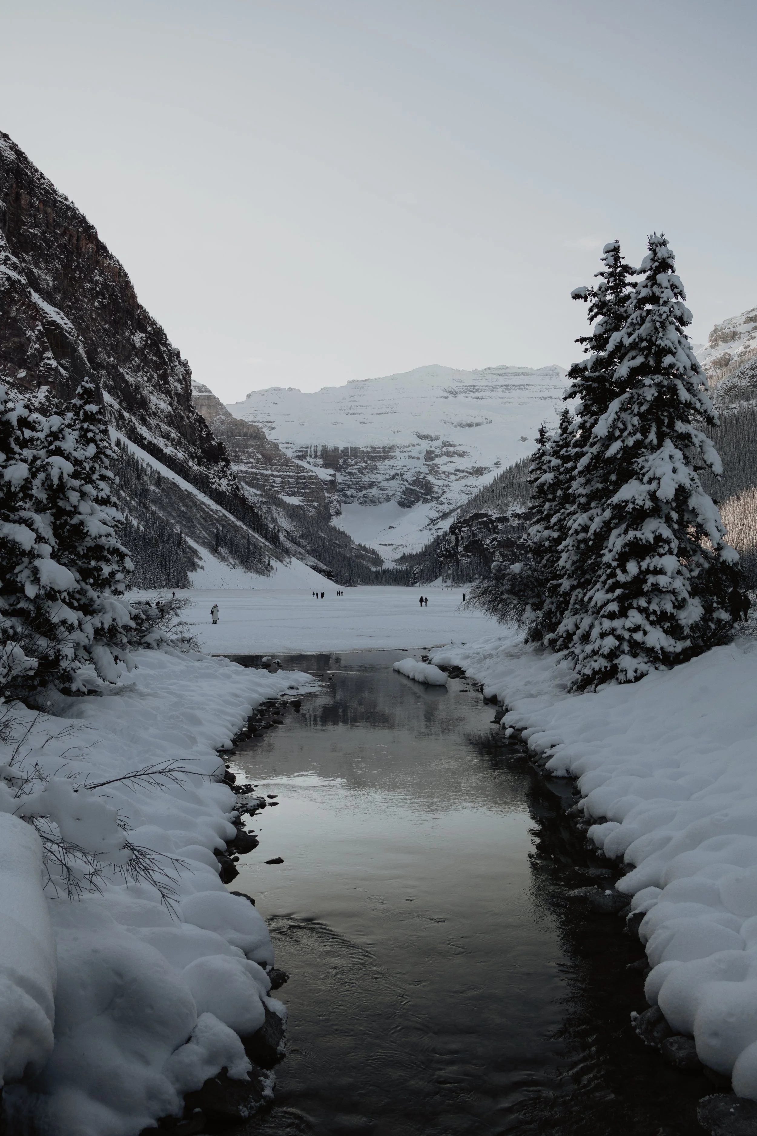 Snow-covered trees and mountains surrounding a partly frozen river, with a snowy lake and distant hikers in a winter landscape.