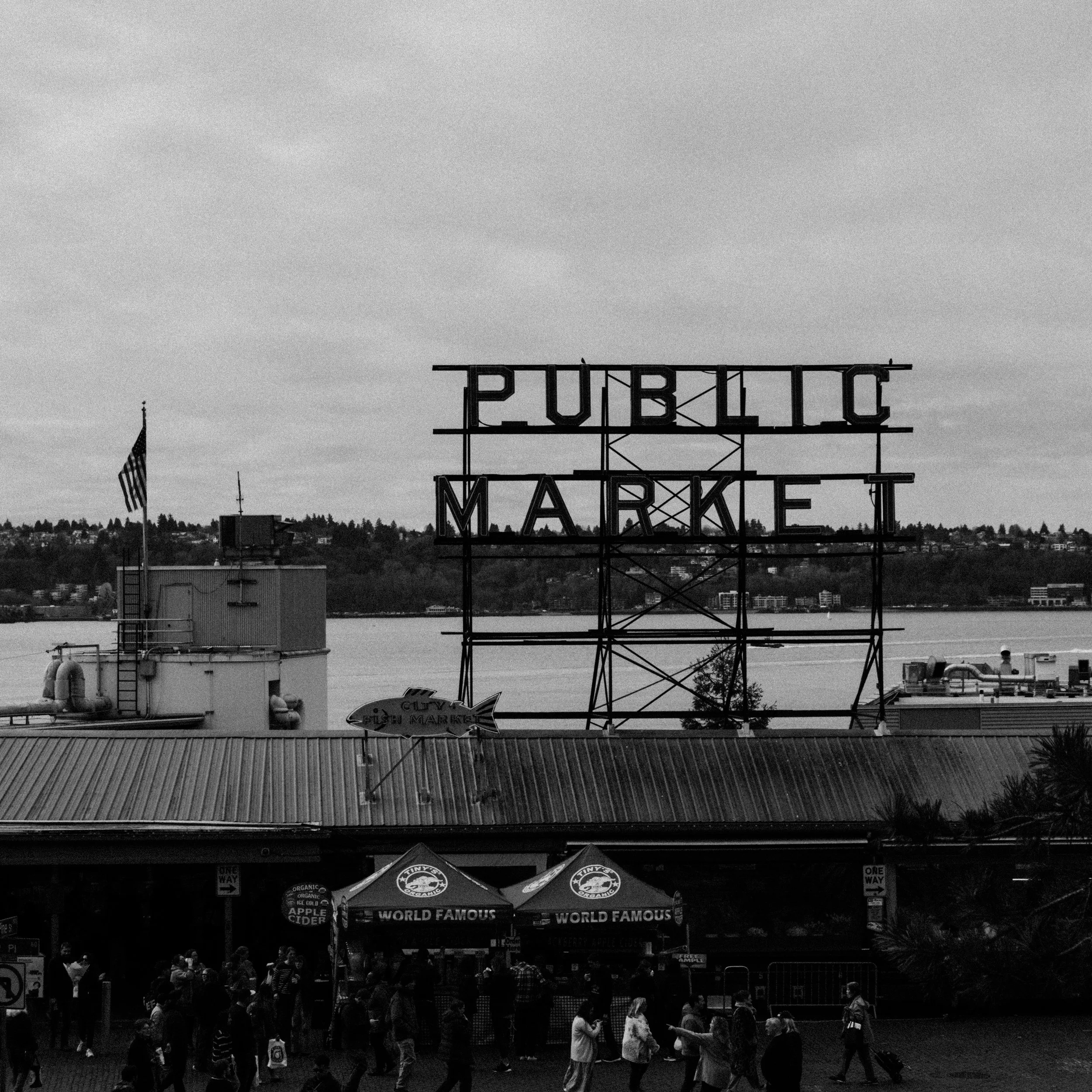 Black and white photo of a public market with a large neon sign that reads 'PUBLIC MARKET'. People are gathered below the sign, with some waiting in line at a food stand with umbrellas that say 'WORLD FAMOUS'. An American flag is flying on a pole to 