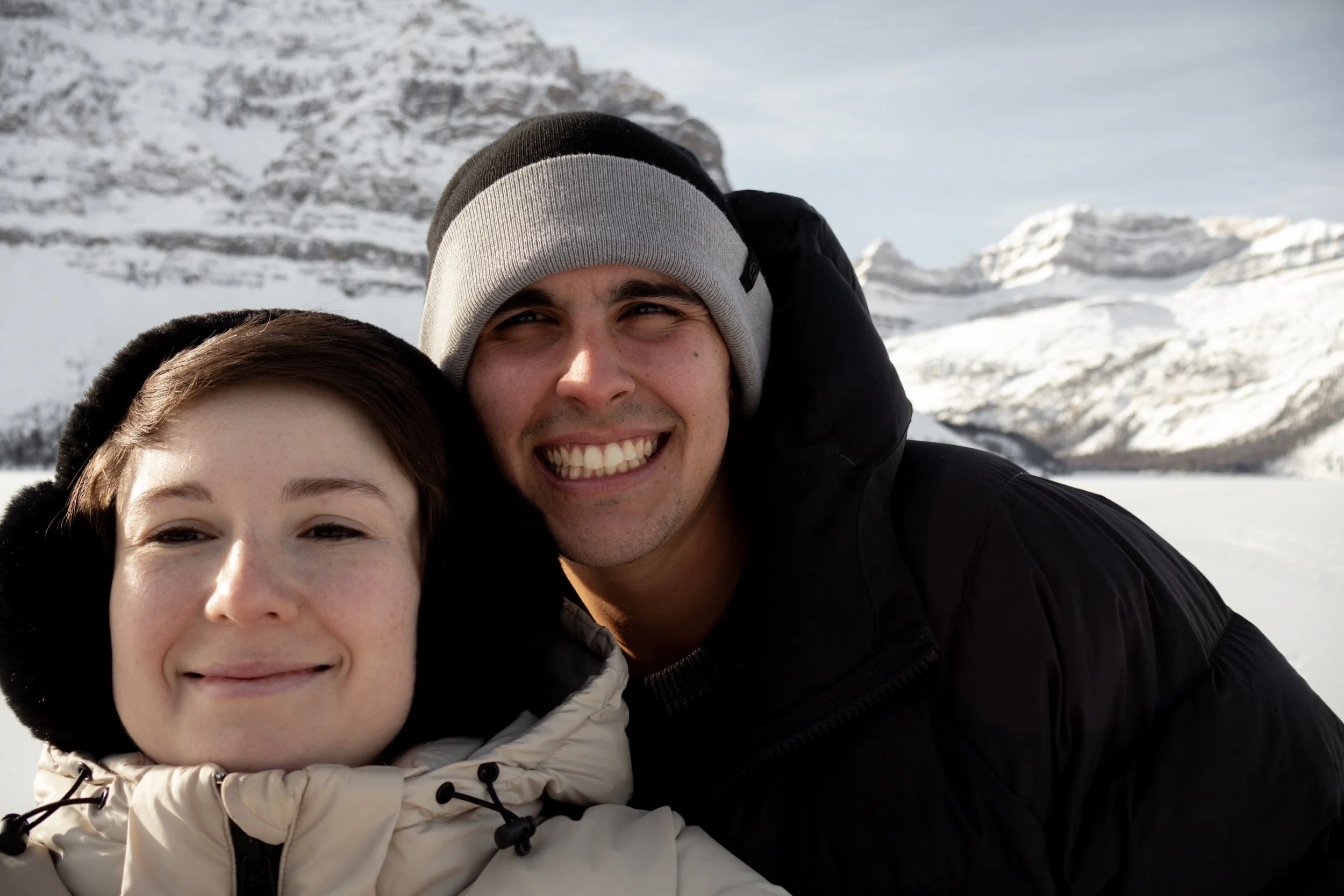 A smiling couple taking a selfie in a snowy mountain landscape, dressed in winter clothing.