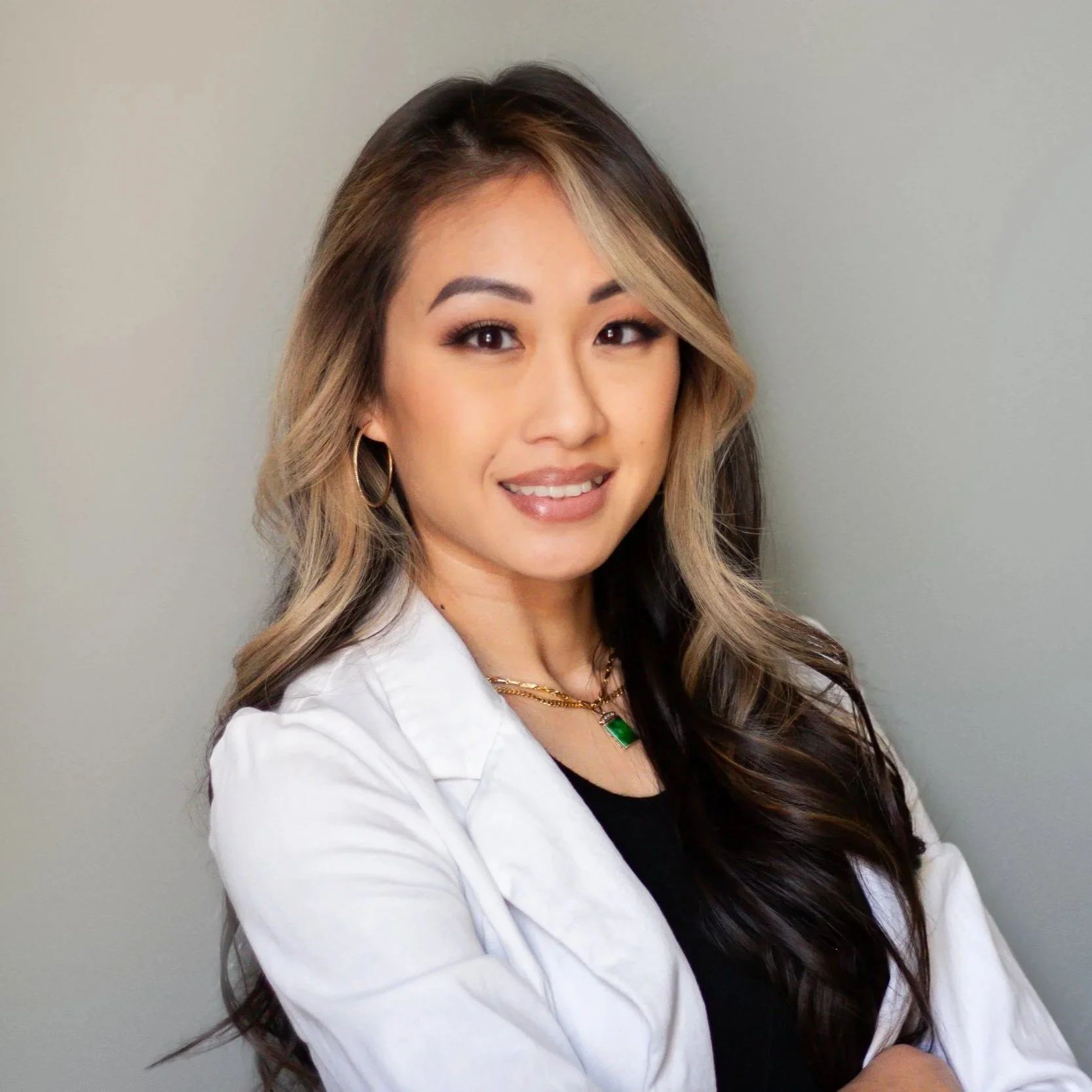 Professional headshot of a woman with long, dark hair, wearing a white blazer, gold jewelry, and smiling against a plain gray background.
