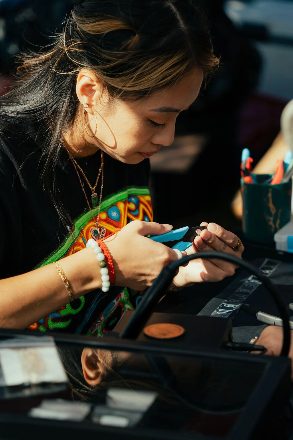 A young woman with blonde highlights is looking down and working with her jewelry pliers, wearing gold jewelry and a colorful shirt, with various objects on the table in front of her.
