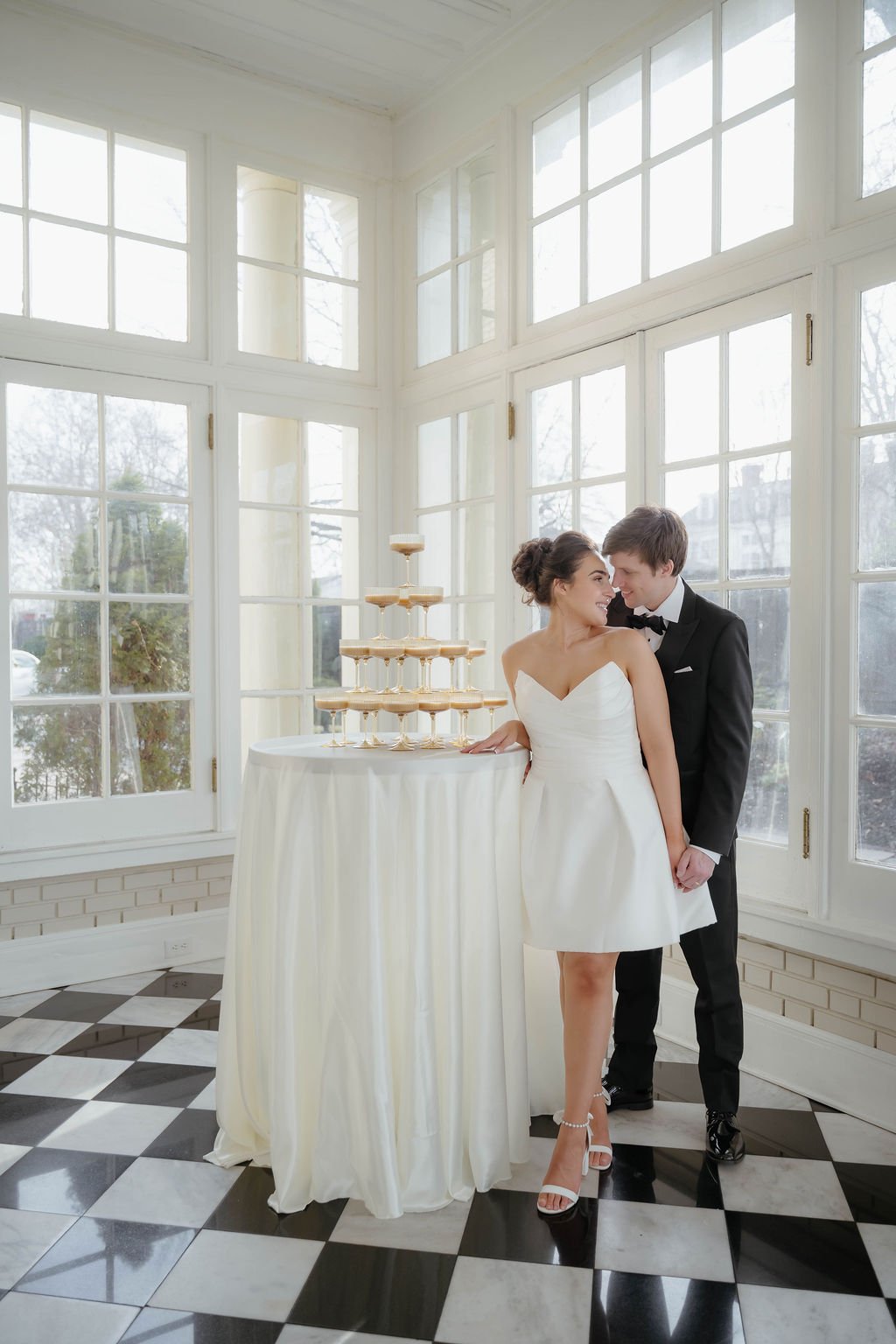 A bride and groom smiling together at a wedding reception next to a champagne tower on a round table, inside a bright room with large windows.