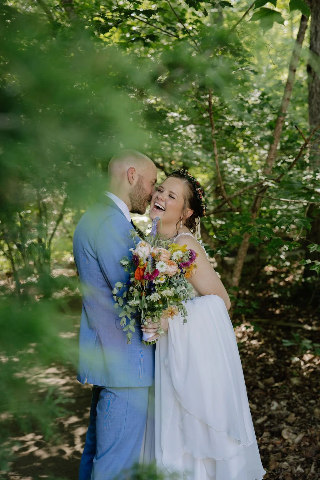 A newlywed couple in wedding attire standing close together in a forest near Charlotte, smiling and leaning towards each other, with the woman holding a large colorful bouquet of flowers.
