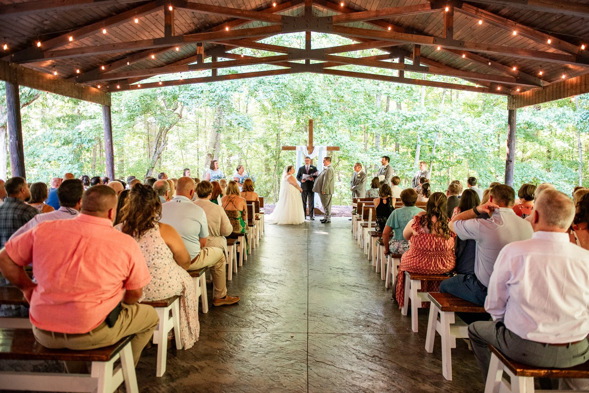 Rustic outdoor chapel ceremony surrounded by lush greenery, with guests seated under wooden beams and string lights as the couple exchanges vows — coordinated by Charlotte Wedding Coordinator.