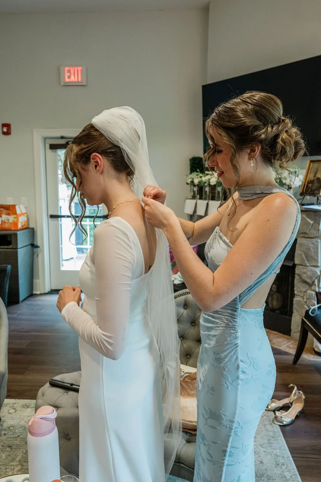 The bride is getting ready with a friend, fastening a borrowed necklace, representing a meaningful ‘something borrowed’ wedding detail symbolising love and support.