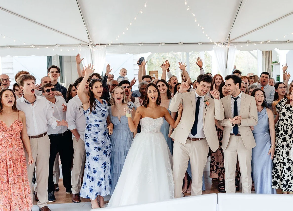 Bride and groom celebrate with guests under a white tent filled with laughter, cheers, and string lights — coordinated by Charlotte Wedding Coordinator.