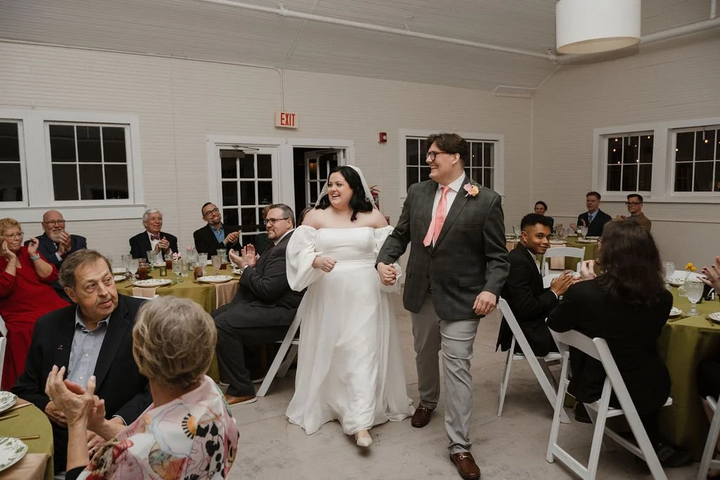 Newlyweds enter their reception hand in hand, smiling as guests applaud and celebrate their arrival — coordinated by Charlotte Wedding Coordinator at Anne Springs Close Greenway.