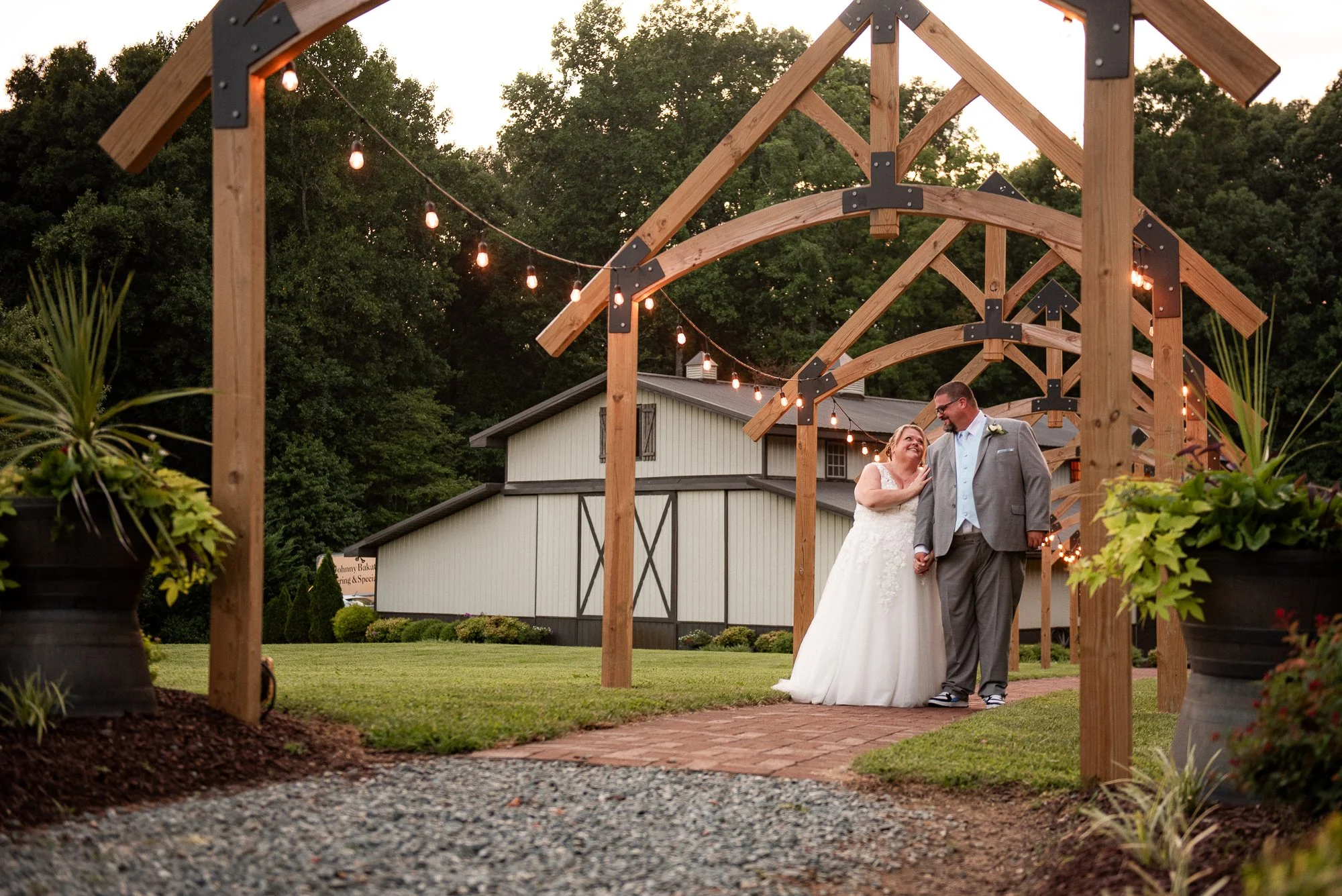 A couple walking under a beautiful outdoor wedding arch with string lights on a summer evening.