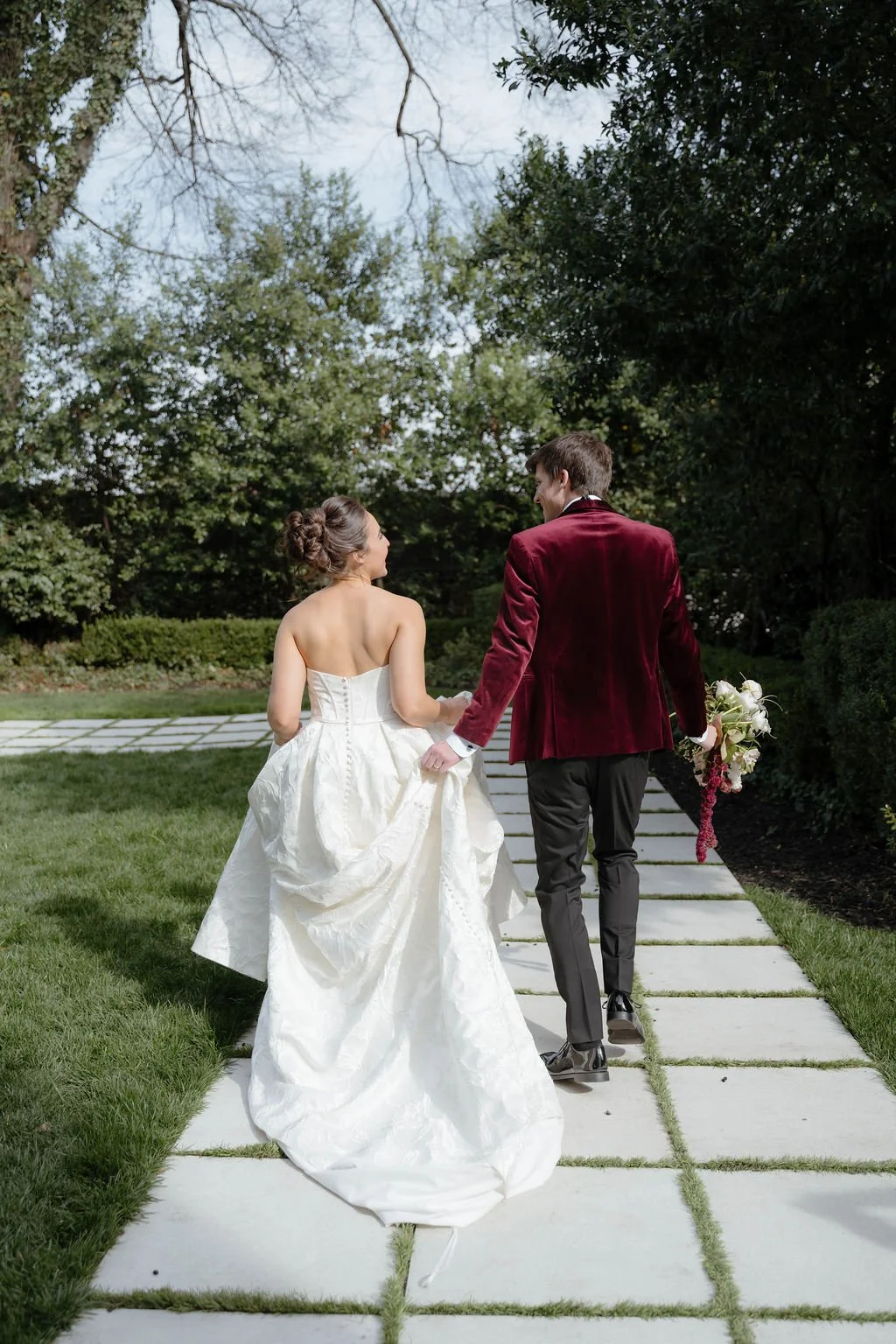 A bride and groom walking hand in hand outdoors on a paved path surrounded by greenery, with the bride wearing a white strapless wedding gown and the groom in a burgundy velvet jacket and black pants, holding a bouquet of flowers.