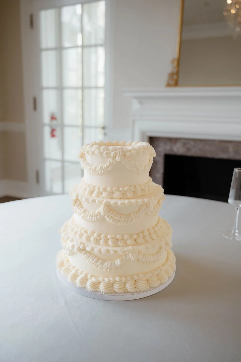 A tall, four-tiered white wedding cake with piped frosting, placed on a white tablecloth in a room with a fireplace and a windowed door in the background.