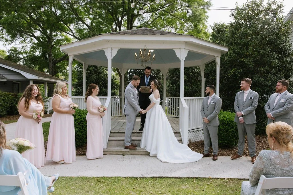 Outdoor gazebo ceremony with the couple holding hands as bridesmaids in blush dresses and groomsmen in gray suits look on — planned by Charlotte Wedding Planner.