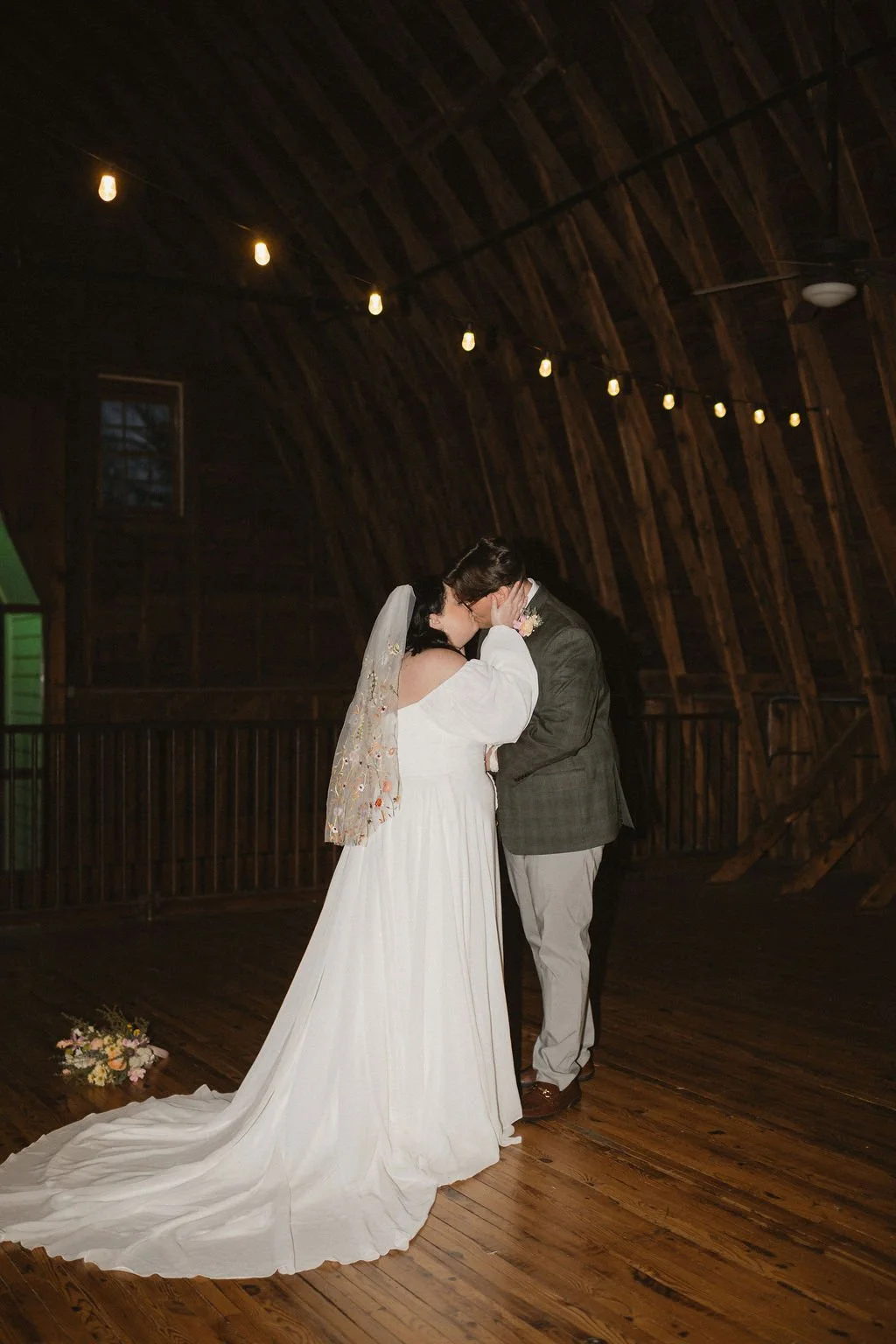 Bride and groom share a kiss under soft string lights in a rustic barn setting, surrounded by warm wood tones and romantic ambiance — coordinated by Charlotte Wedding Coordinator at Anne Springs Close Greenway.