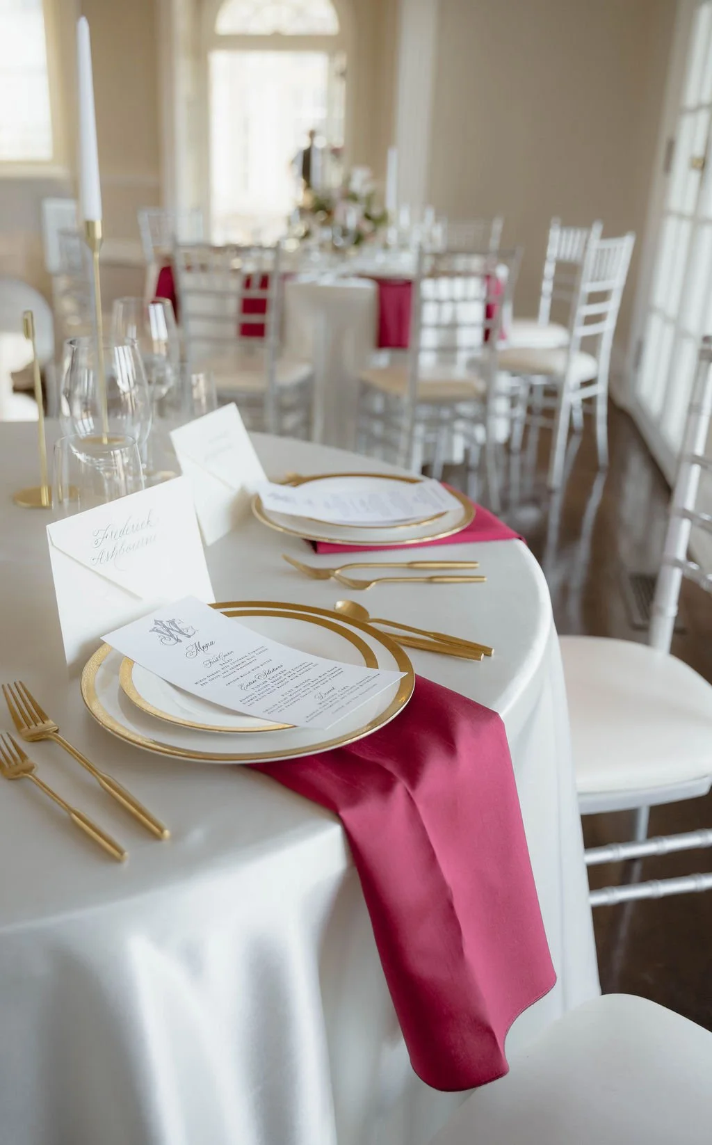 Elegant wedding reception table setup with white tablecloth, gold cutlery, red and white napkins, and printed menus. The background shows more tables with similar arrangements and white chairs.