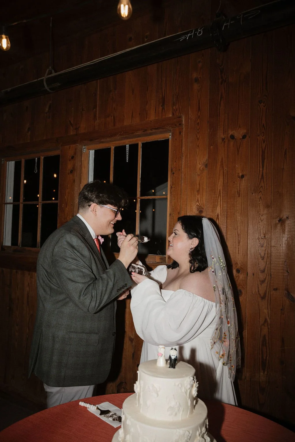 Couple shares a sweet moment feeding each other cake beside a three-tiered white wedding cake with floral details — coordinated by Charlotte Wedding Coordinator